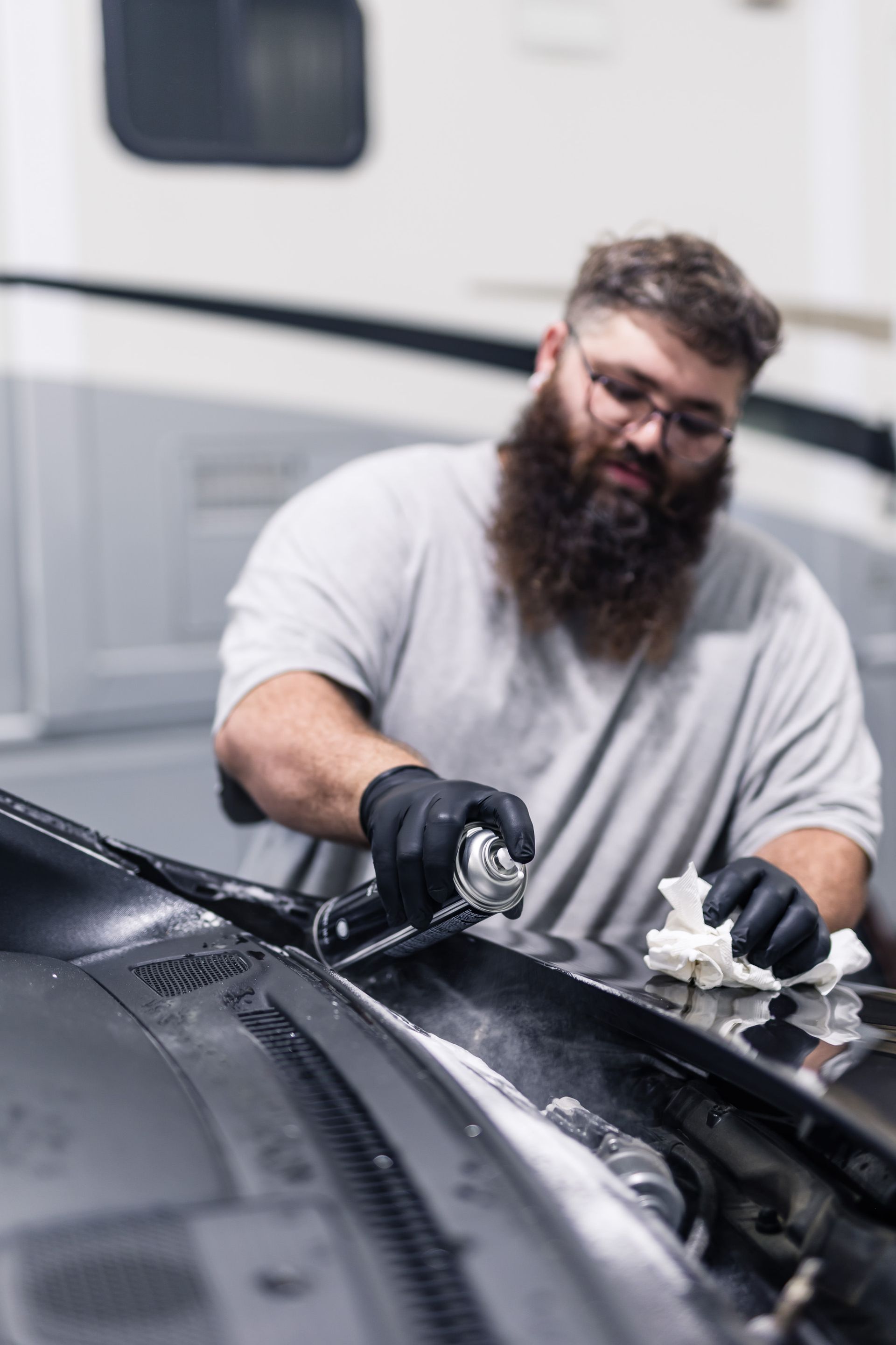 A bearded man wearing gloves sprays something on a car hood in a shop. He's holding a white cloth in his other hand.