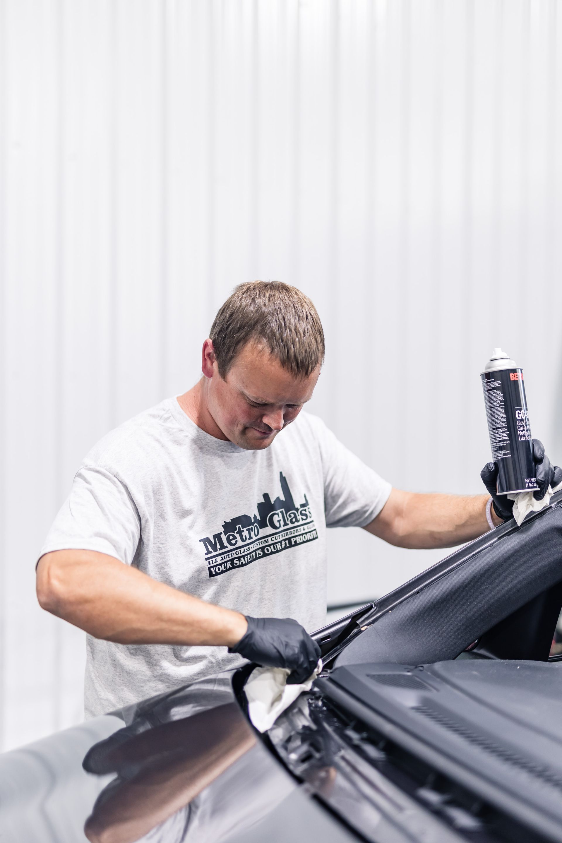 A man in a gray shirt and black gloves applies sealant to a car part indoors. The setting is a well-lit workshop.