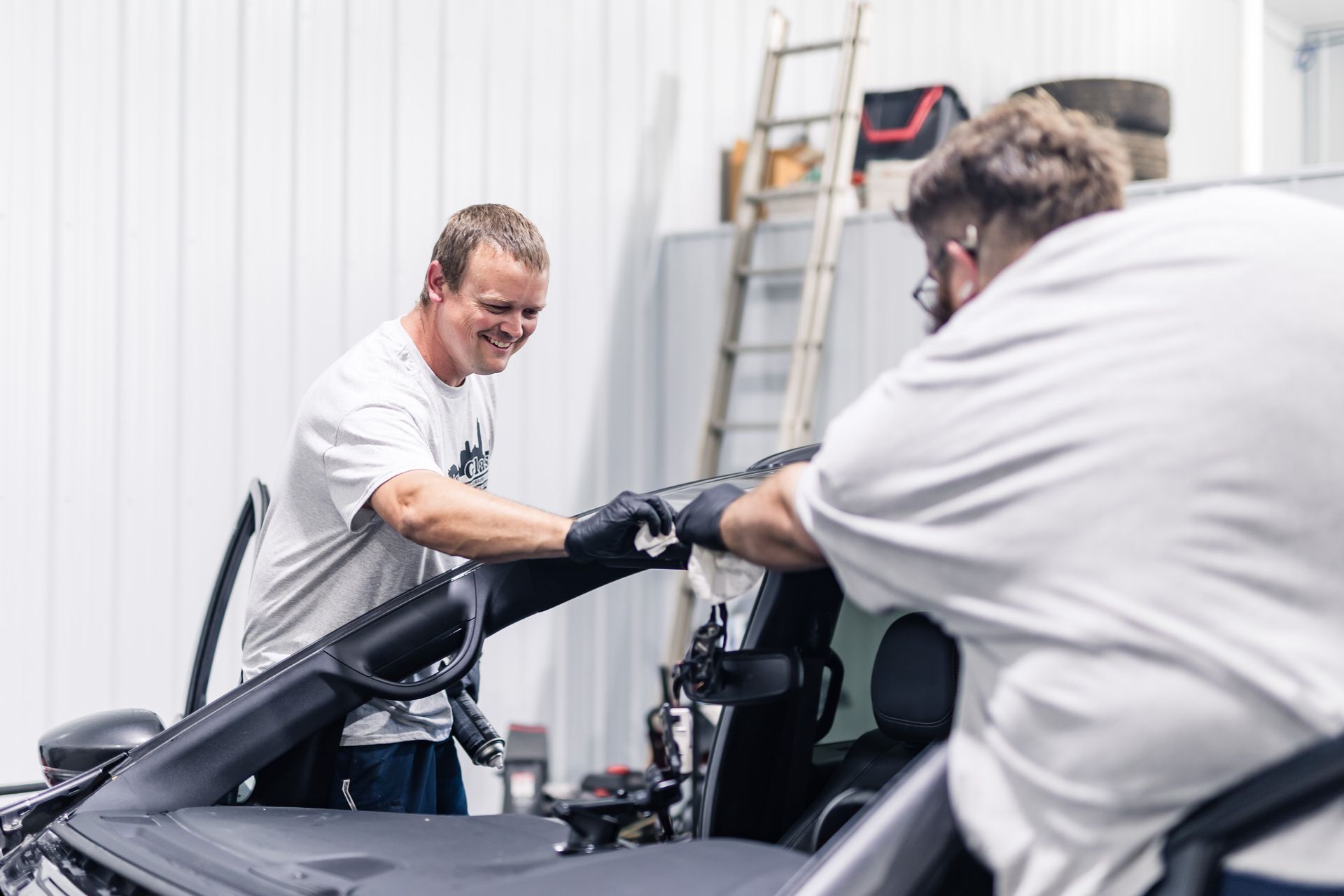 Two men installing a black car soft top in a garage. One man smiles while working, the other is hunched over.