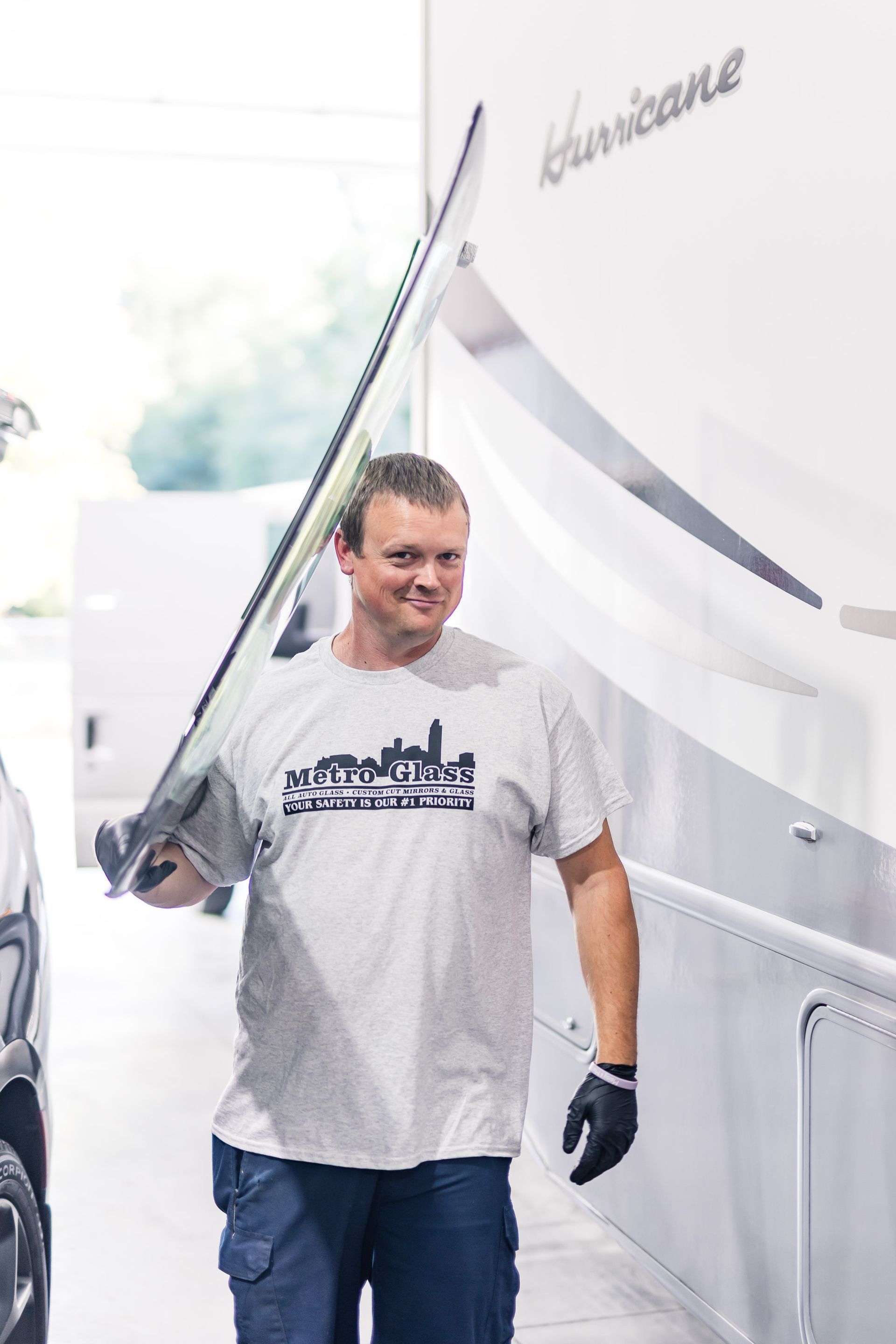 Man in a grey t-shirt carries a long piece of glass near a white RV. He smiles, wearing a black glove on one hand.
