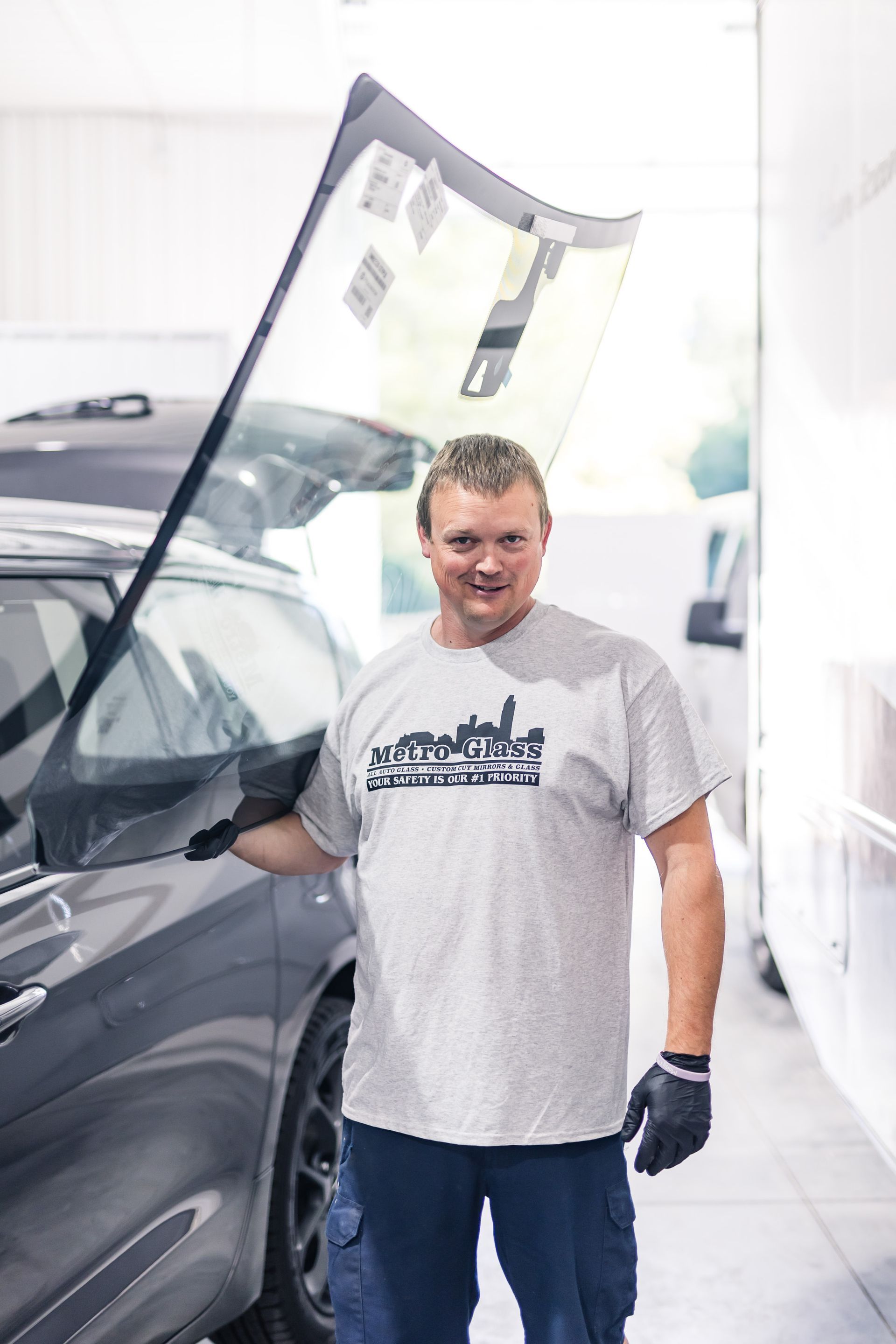 Mechanic holds a windshield, smiling next to a car in a garage. He wears gloves and a gray t-shirt.