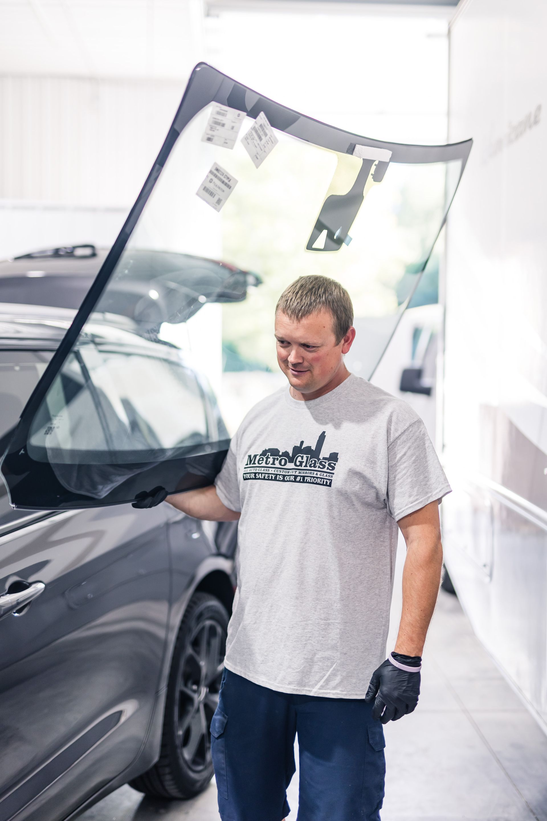 A man holding a car windshield next to a dark gray car. He's wearing a gray shirt, blue pants, and black gloves, smiling in a brightly lit workshop.