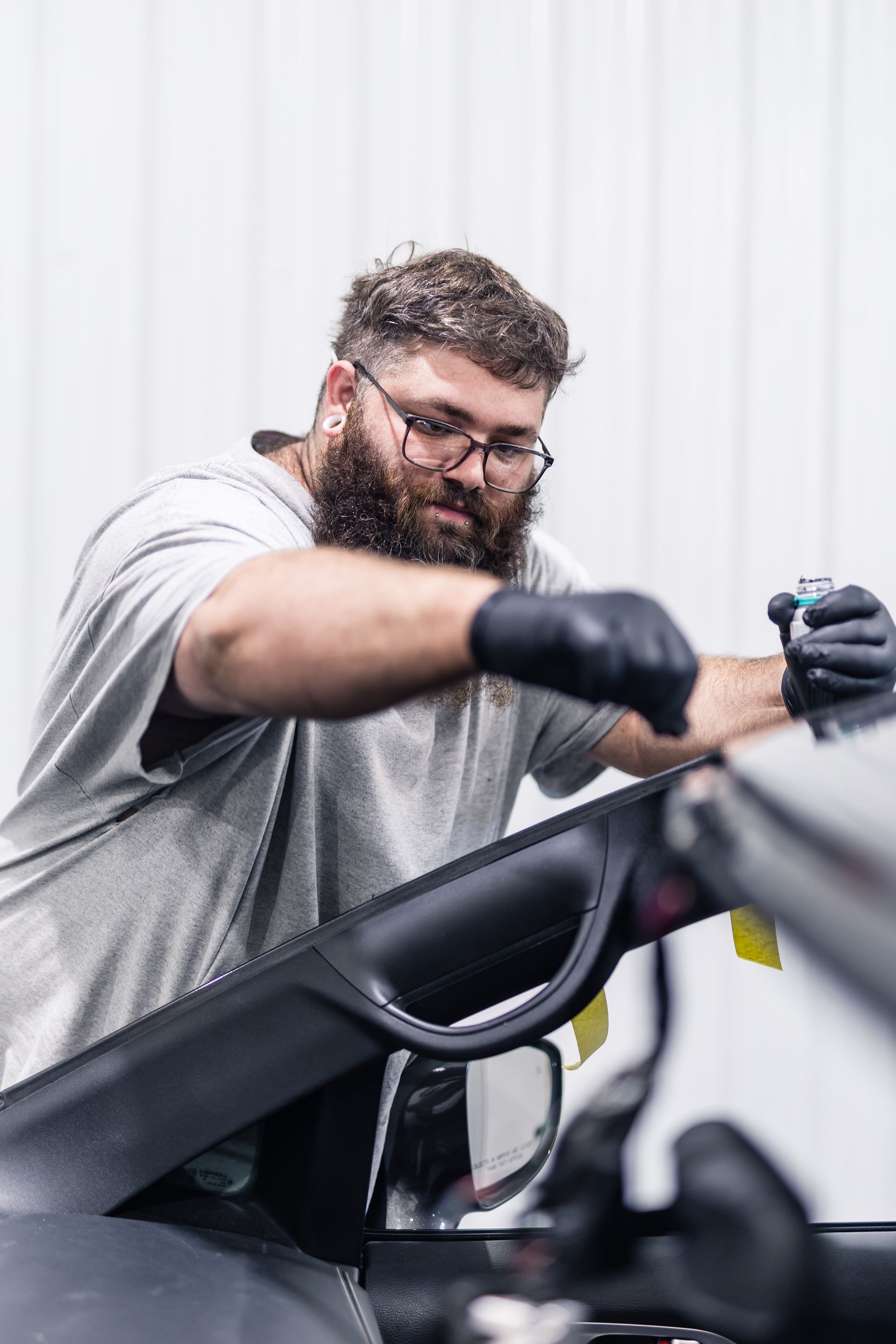 Man in glasses, beard, and black gloves applying product to a car's windshield in a well-lit workshop.