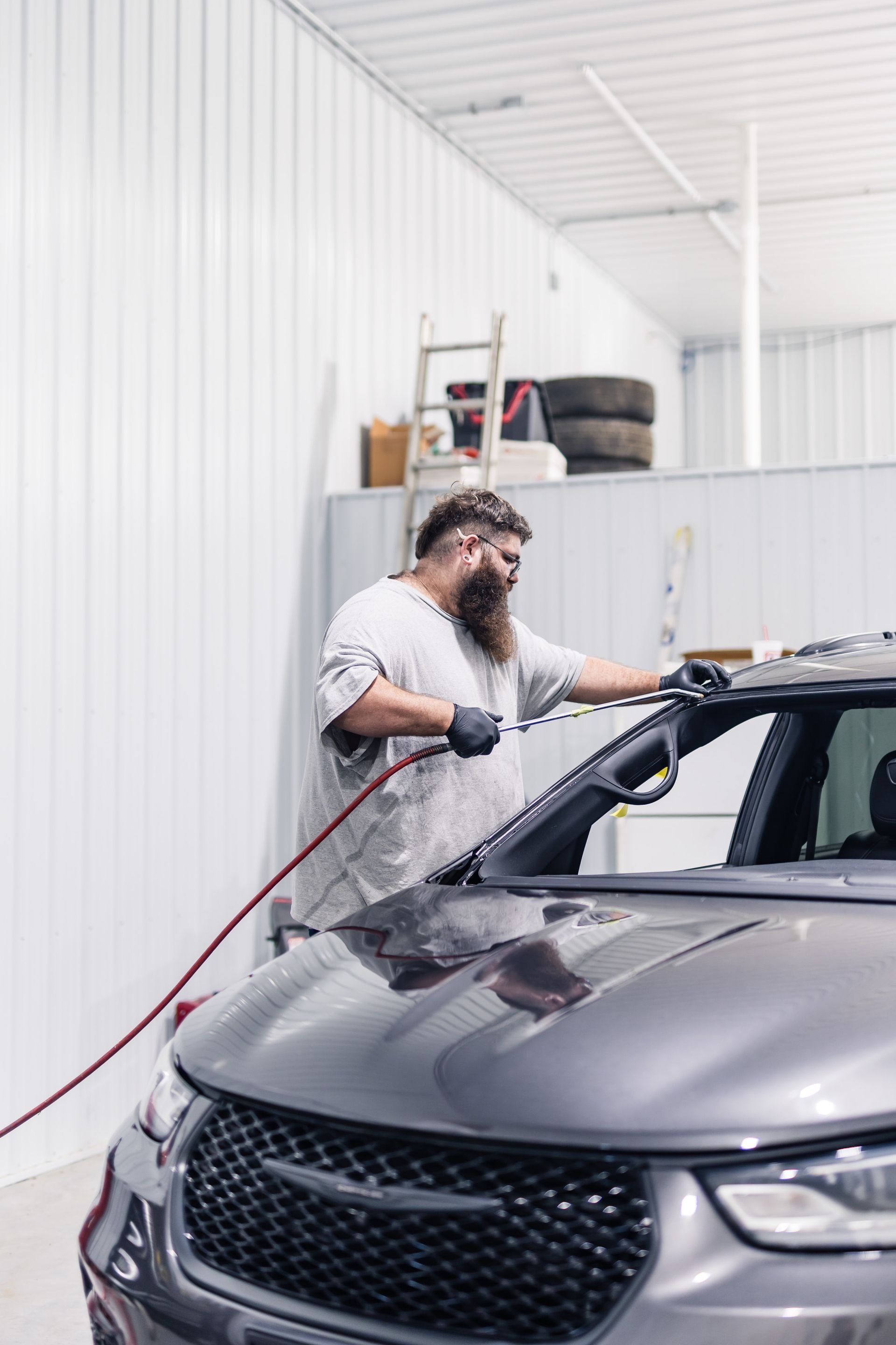 A man washes a gray car in a white-walled garage. He holds a hose and wears gloves.
