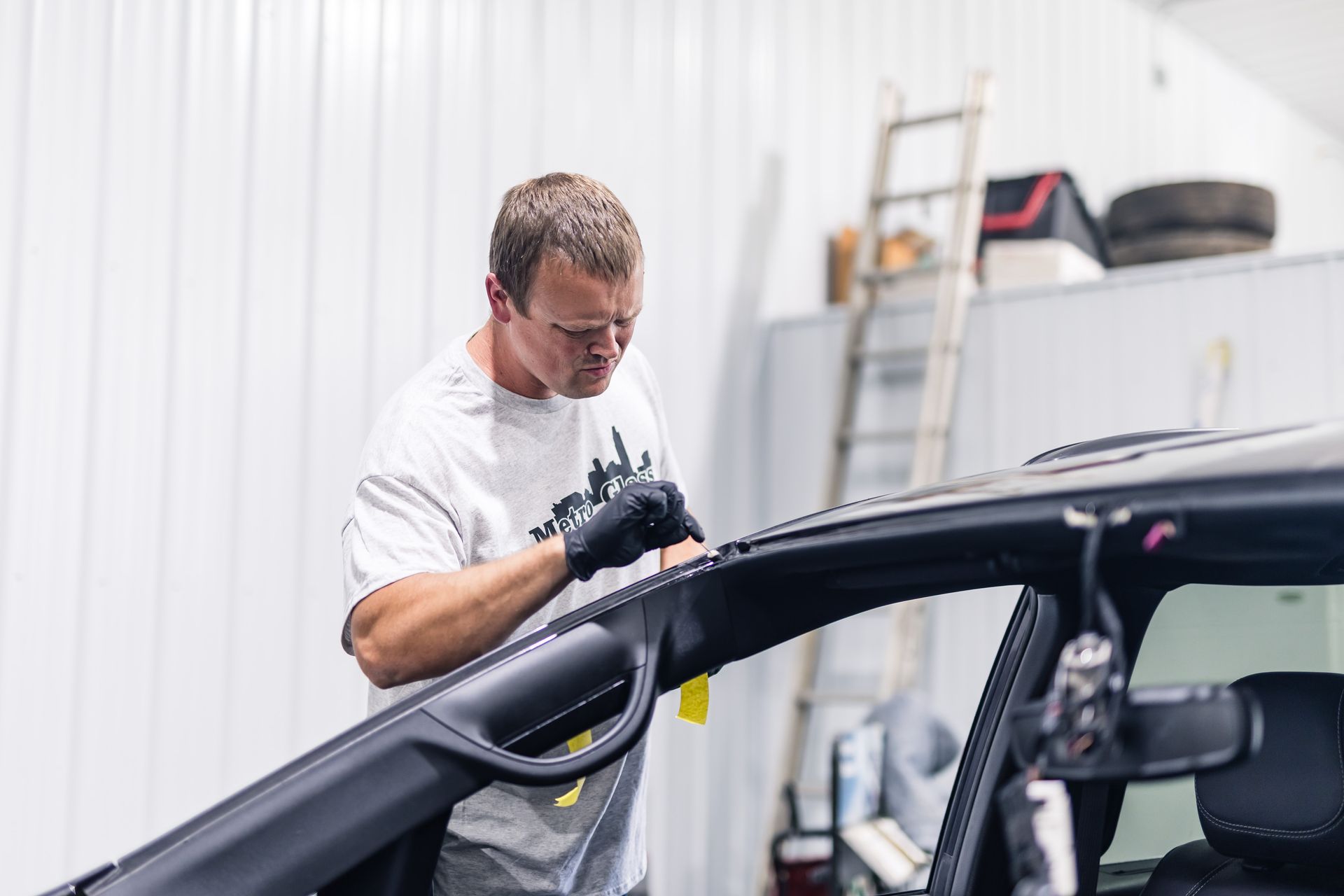 A man in a white shirt and gloves working on a car's black trim inside a brightly lit garage. He's focused and working with precision.