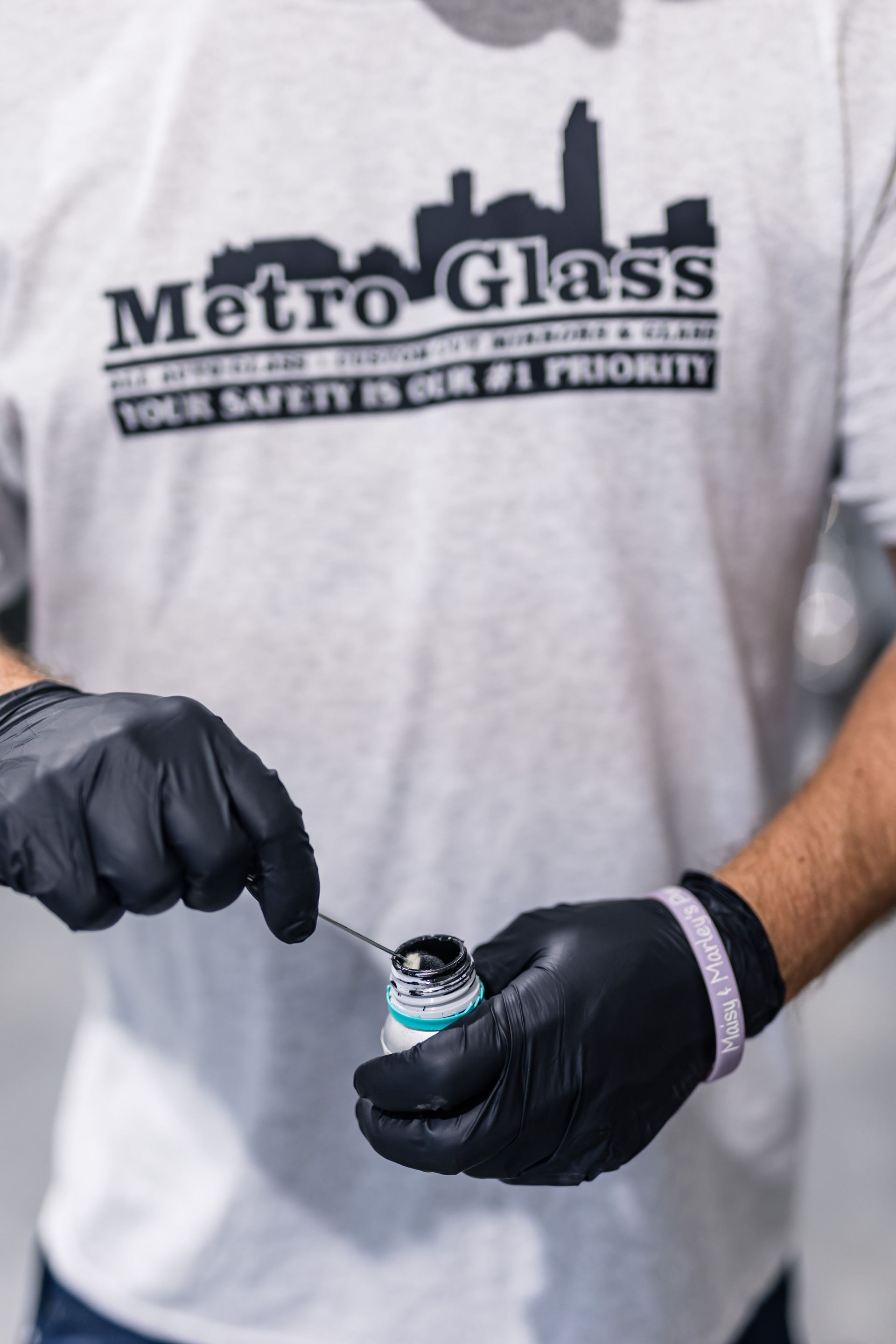 Person in gray t-shirt with Metro Glass logo and black gloves holding a small bottle and tool, likely applying sealant.