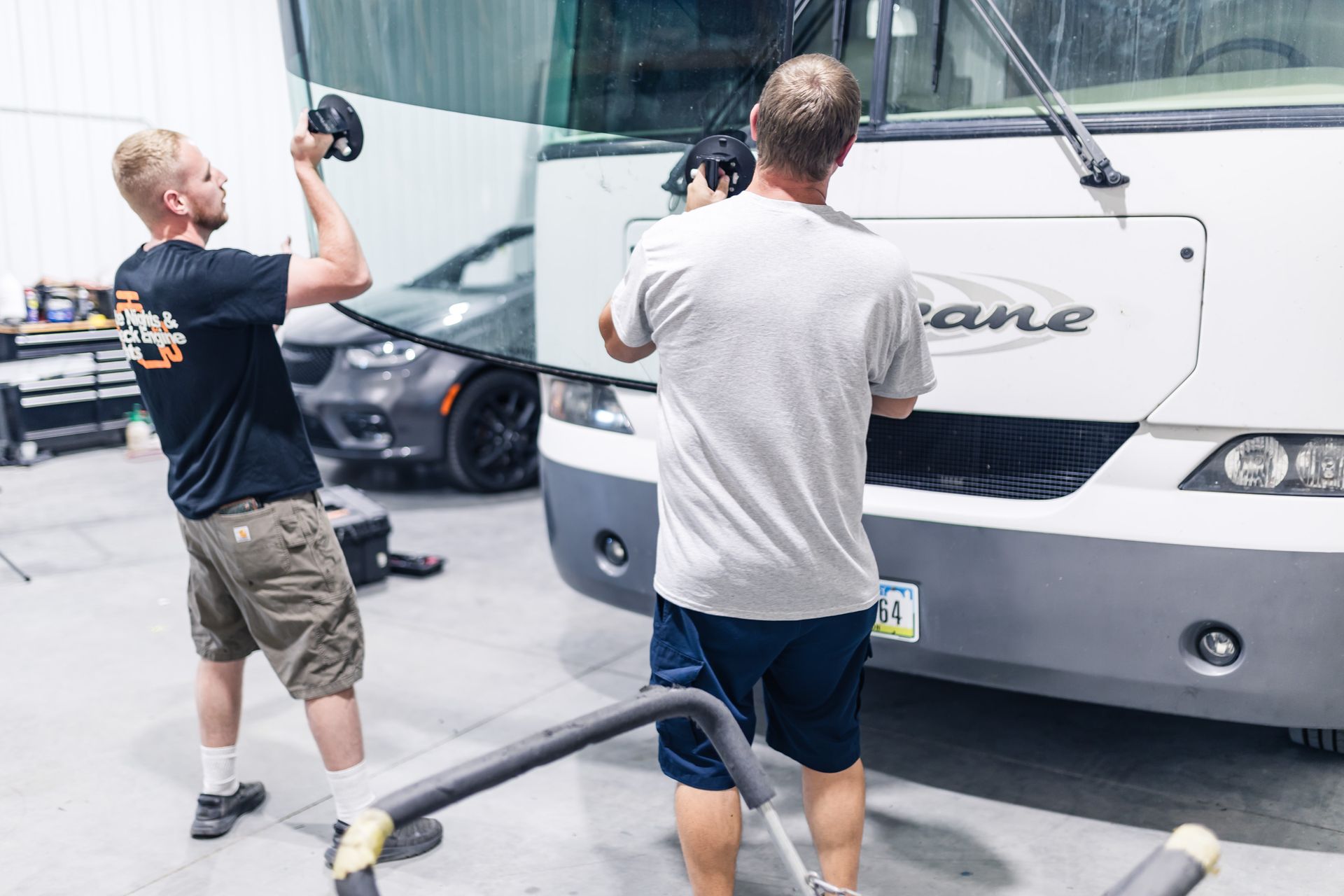 Two men working on the front of a white recreational vehicle inside a garage. One holds a tool overhead, the other uses a tool at the RV.
