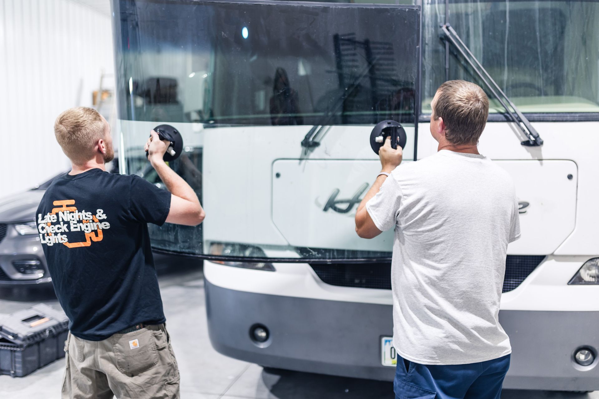 Two men installing a large windshield on a white bus, using suction cups. They are in a workshop.