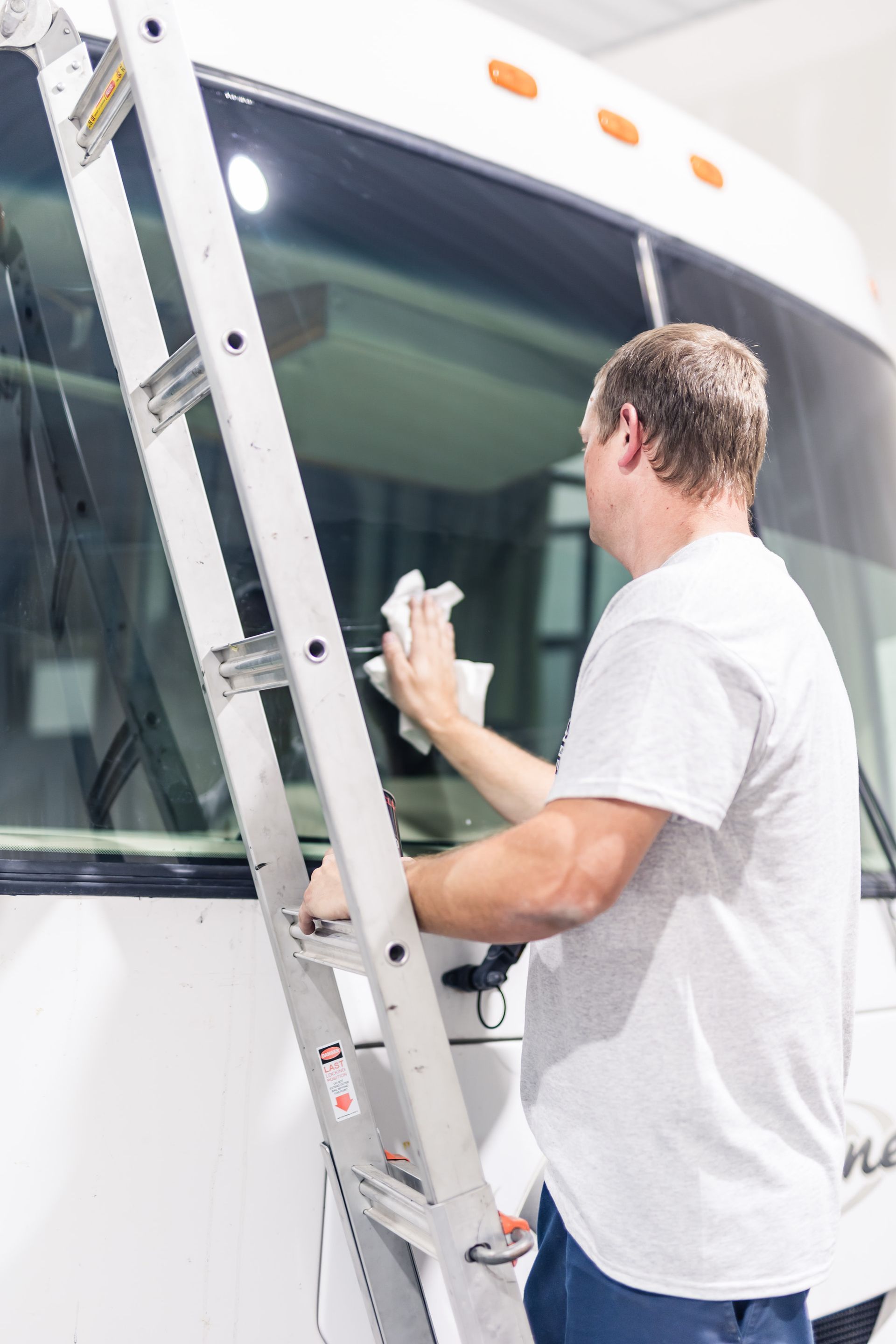 Man cleaning the windshield of a white RV while standing on a ladder.