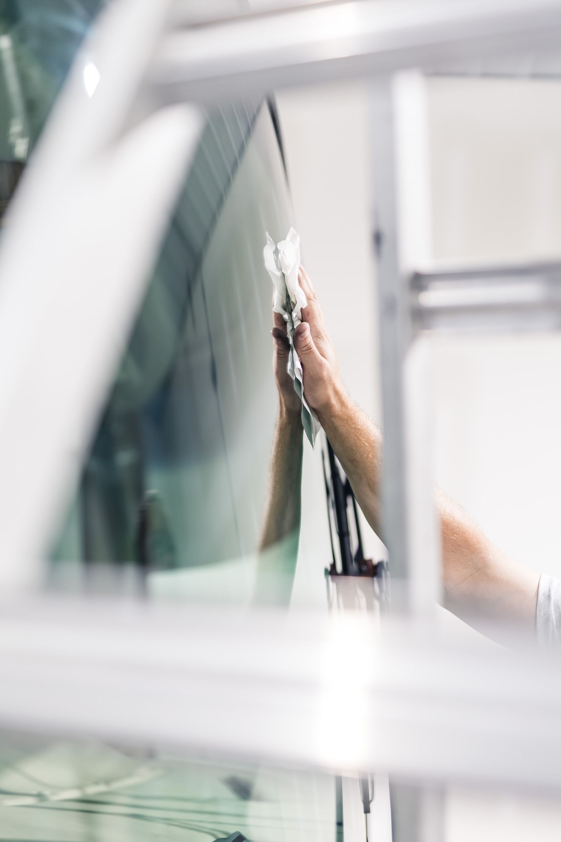 Person applying tint to a car window. A hand holds a tool, smoothing film onto the glass. White and gray tones are present, likely in a workshop.