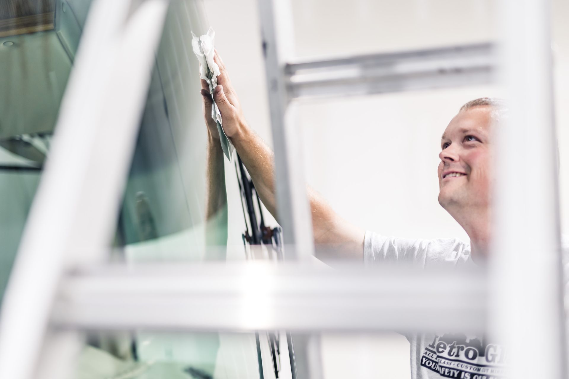 A man on a ladder installing window tint, smiling. He is in a workshop with the window in front of him.
