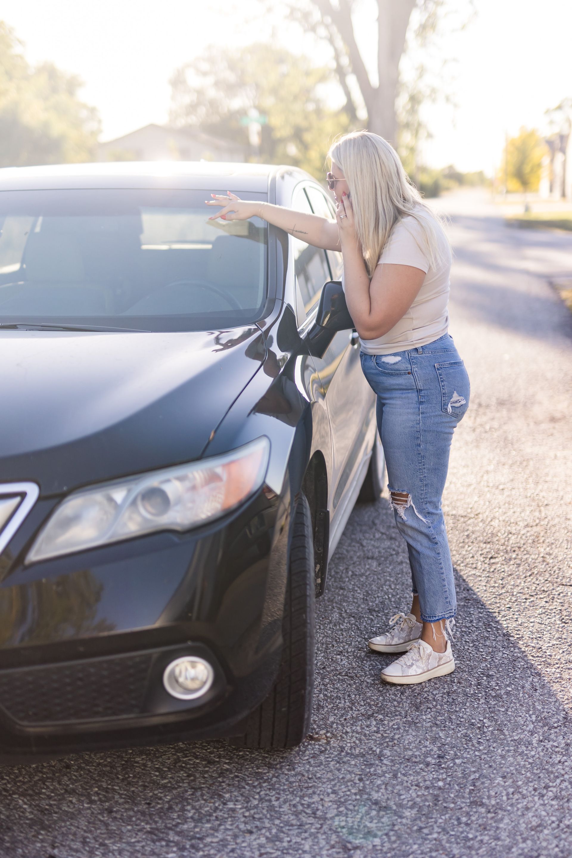 Blonde woman in distressed jeans, leaning against a black car on a street, looking surprised and holding her hand to her face.