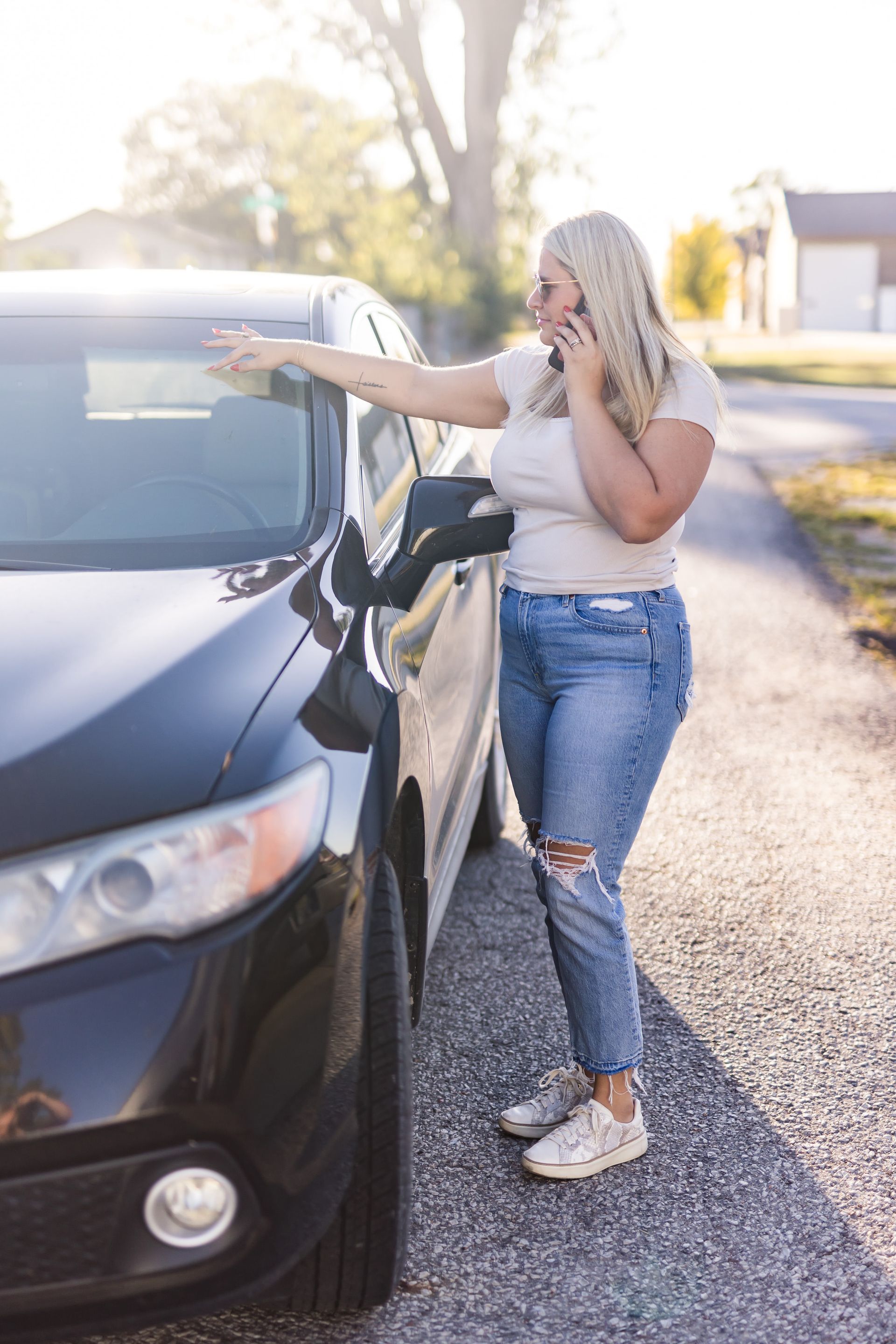 Blonde woman in jeans on phone beside a black car on a sunny driveway. She points toward car window.