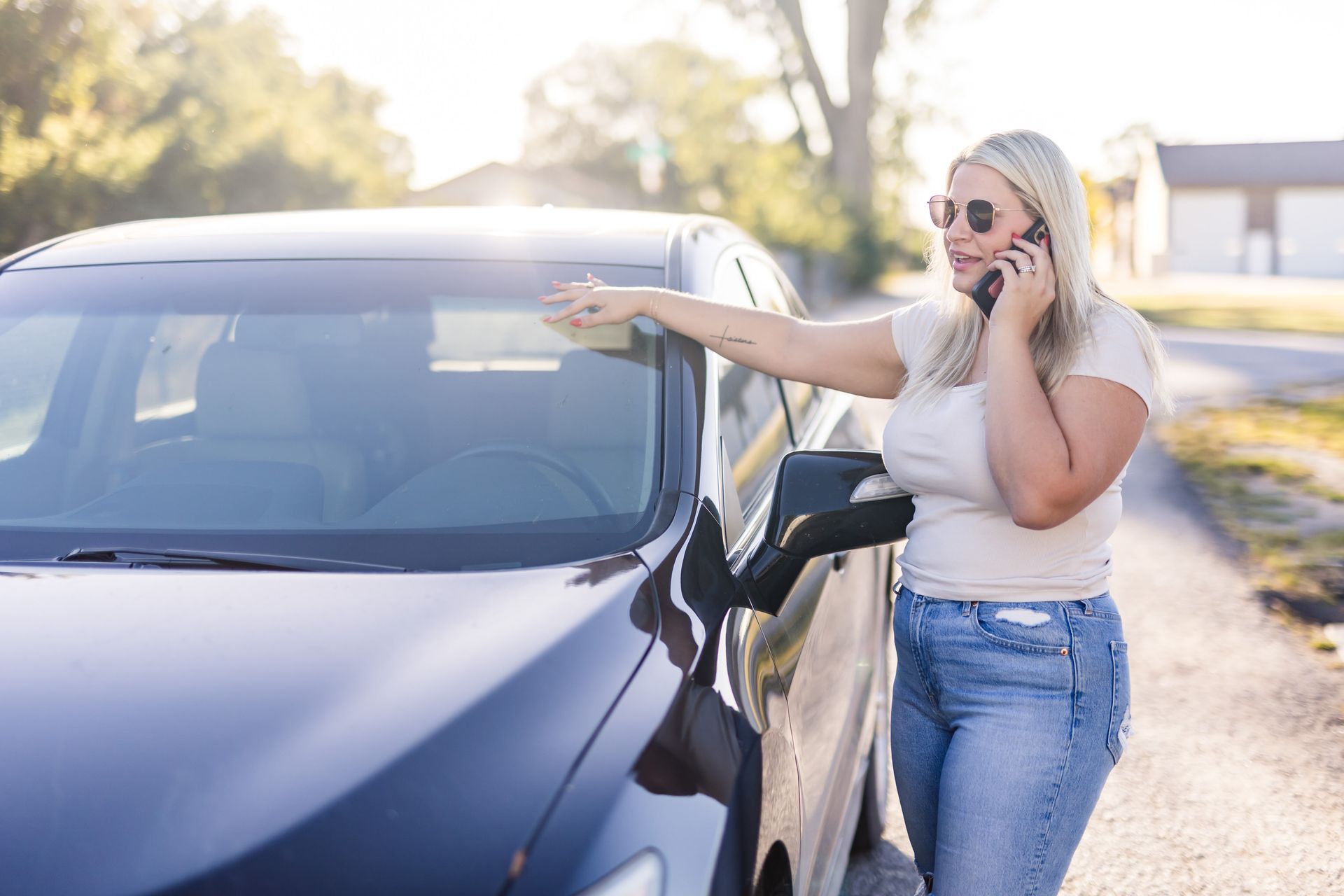 Blonde woman in sunglasses talking on phone, leaning against a dark car on a sunny roadside.