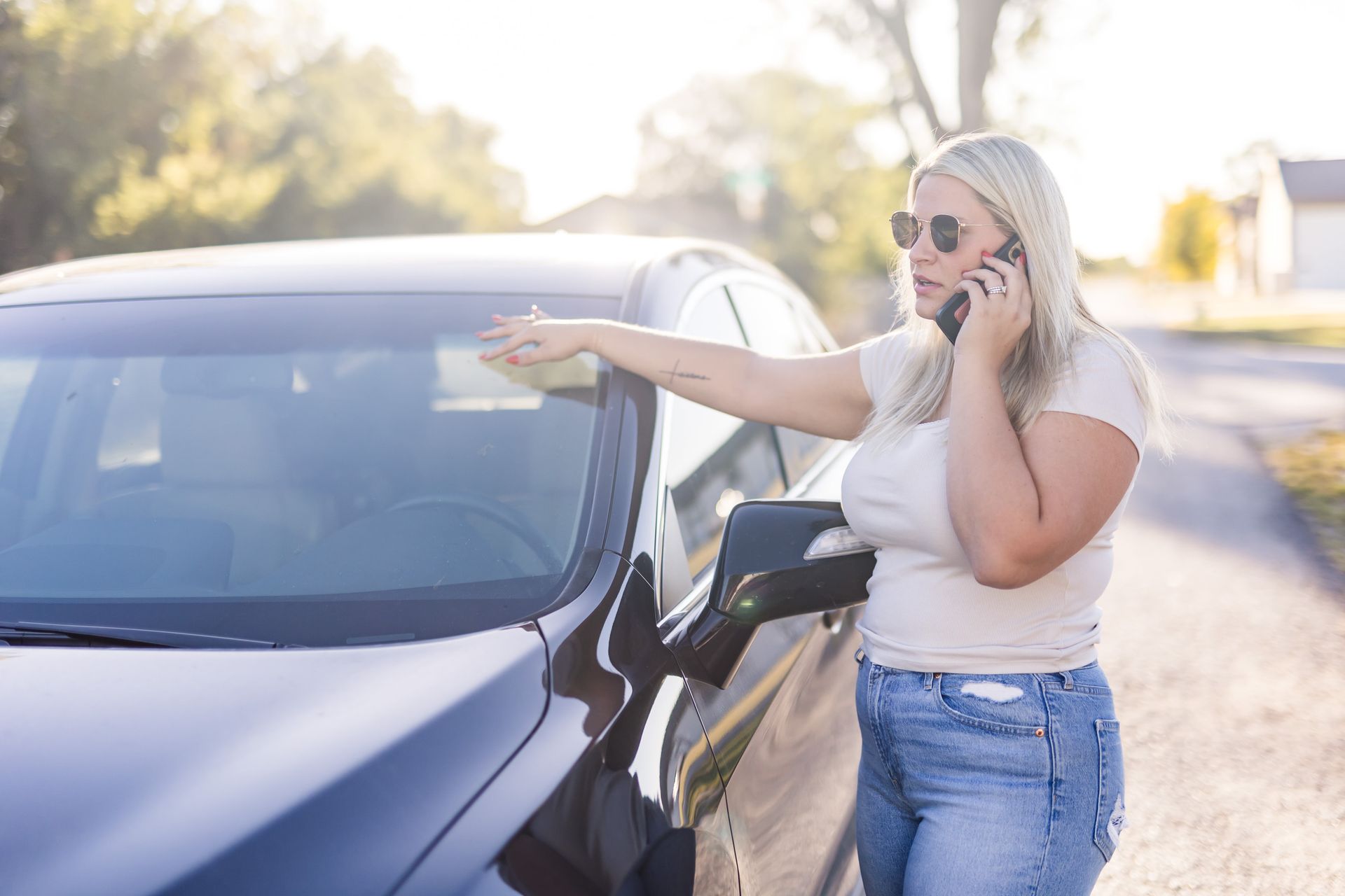 Blonde woman wearing sunglasses on phone next to black car on a rural road, gesturing with her arm.