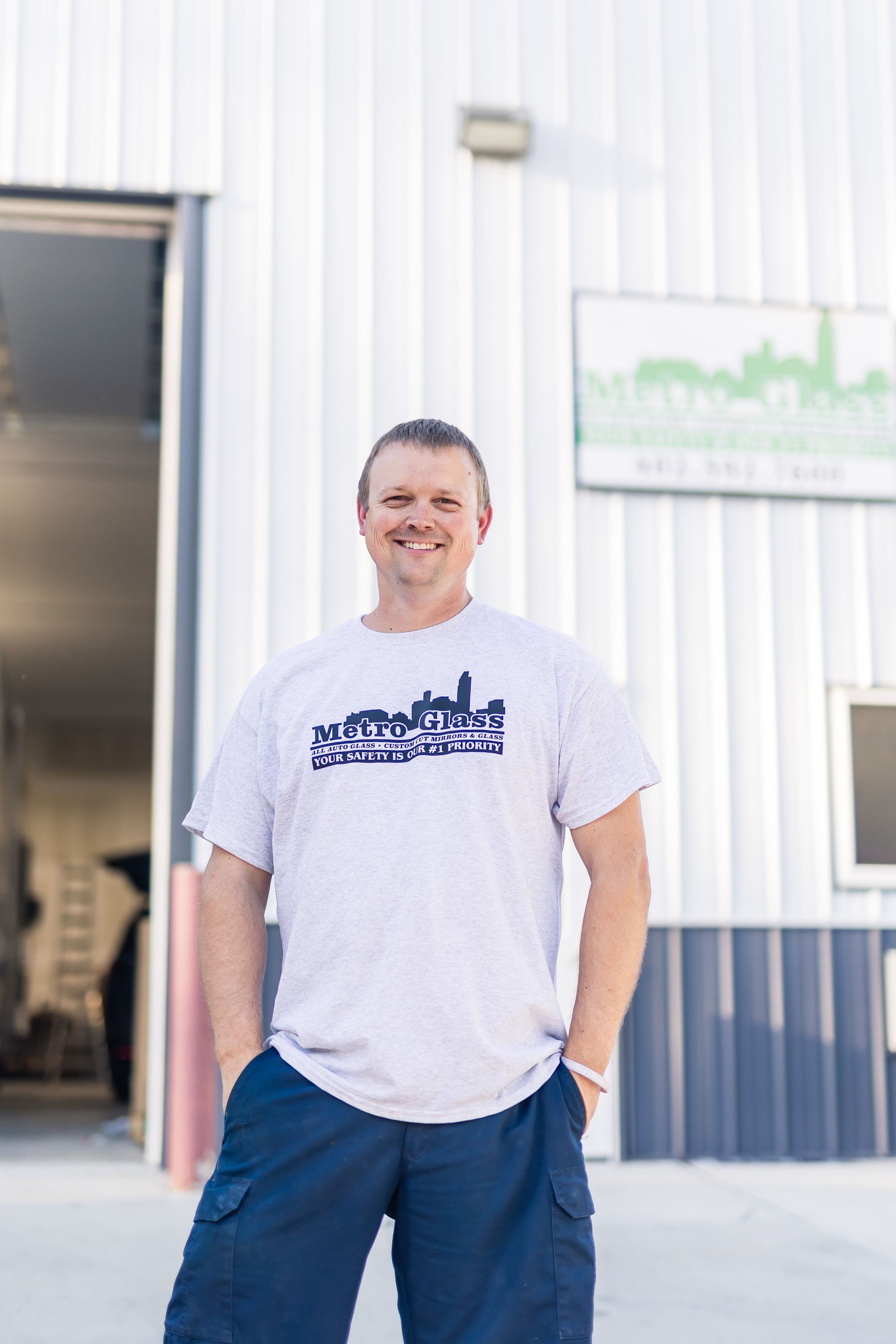 A man in a light gray shirt with a city skyline graphic smiles in front of a building. He has his hands in his pockets.