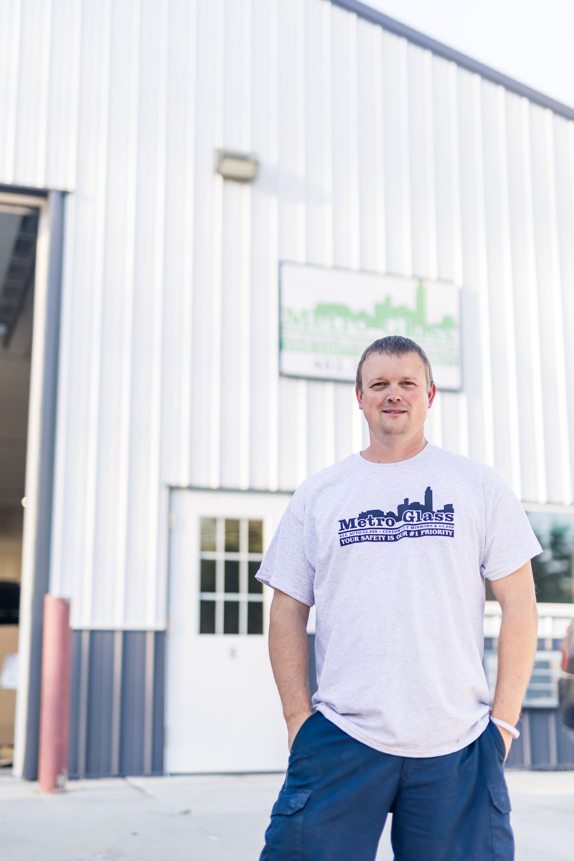 Man in a gray t-shirt stands in front of a white building with a logo, hands in pockets.