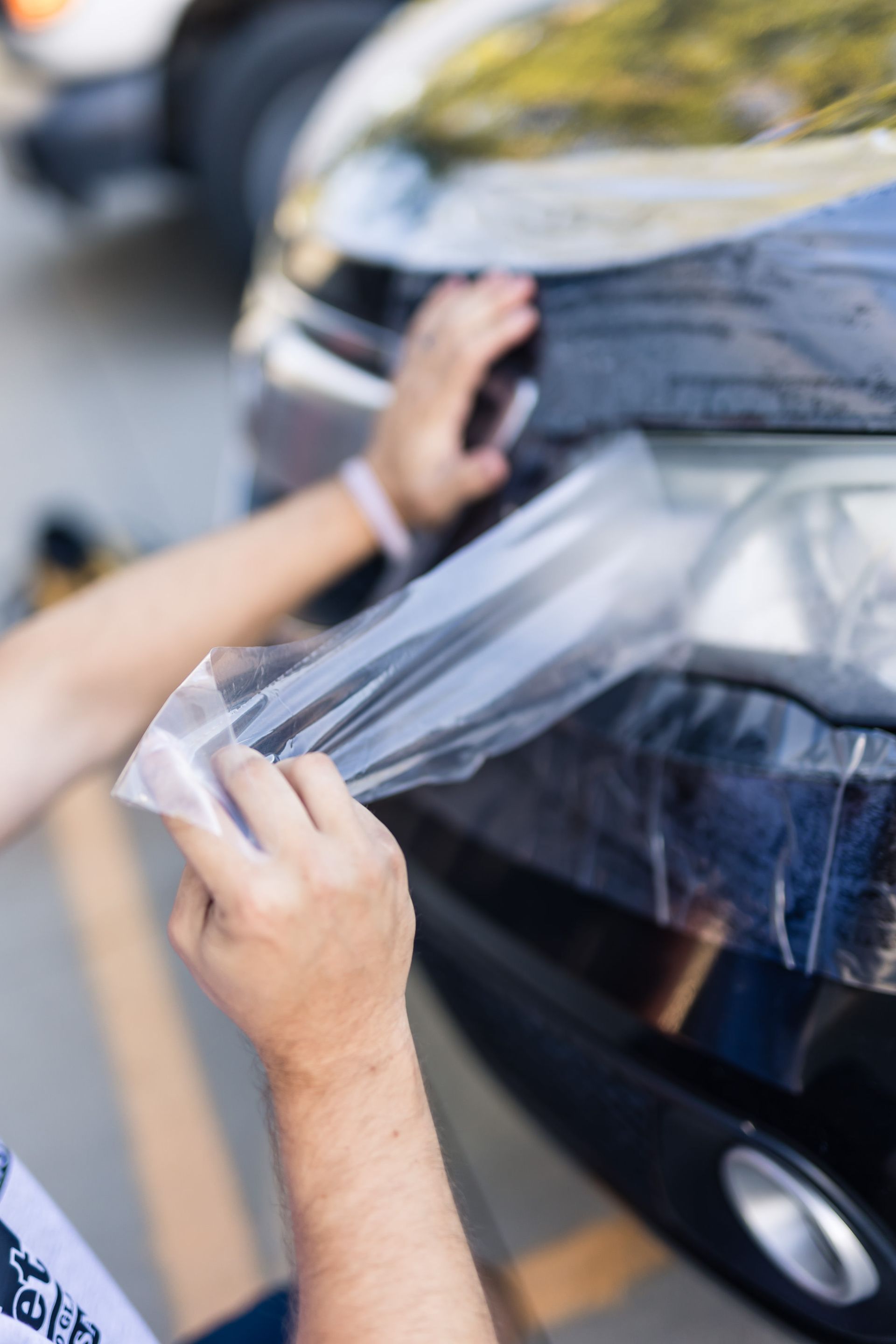 Person applying protective film to the headlight of a black car. They are holding the film with both hands near the car's front.