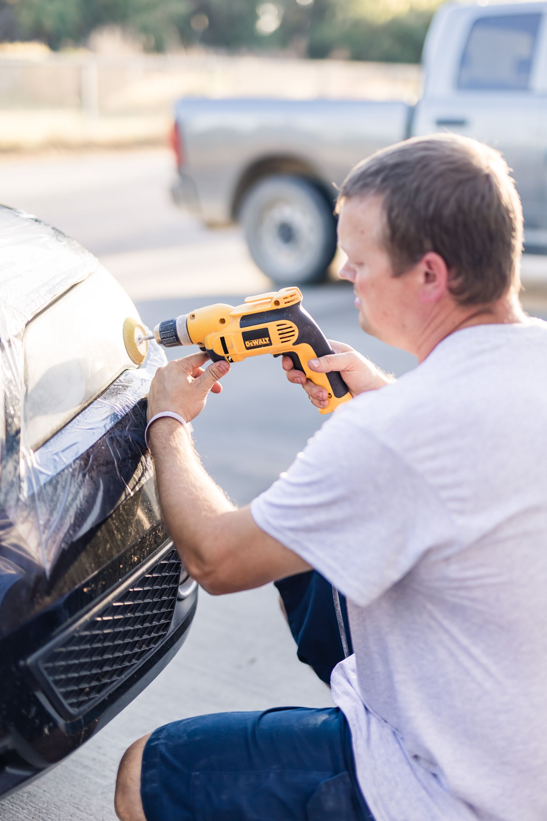Man polishing a black car with a yellow power tool outdoors. He wears a white shirt and kneels near the car.