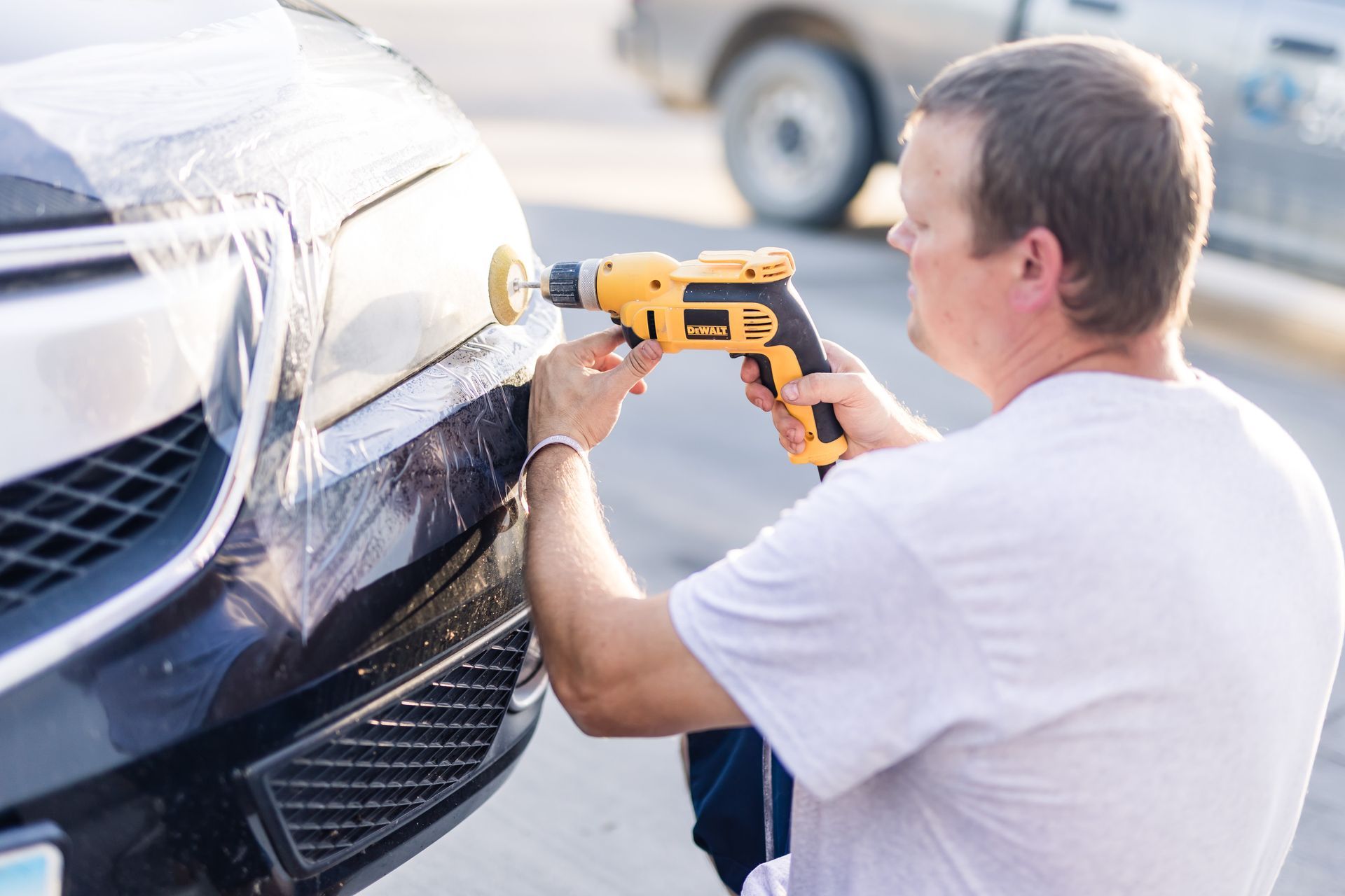 Man using a power drill with a buffing pad to work on the front bumper of a black car outdoors.