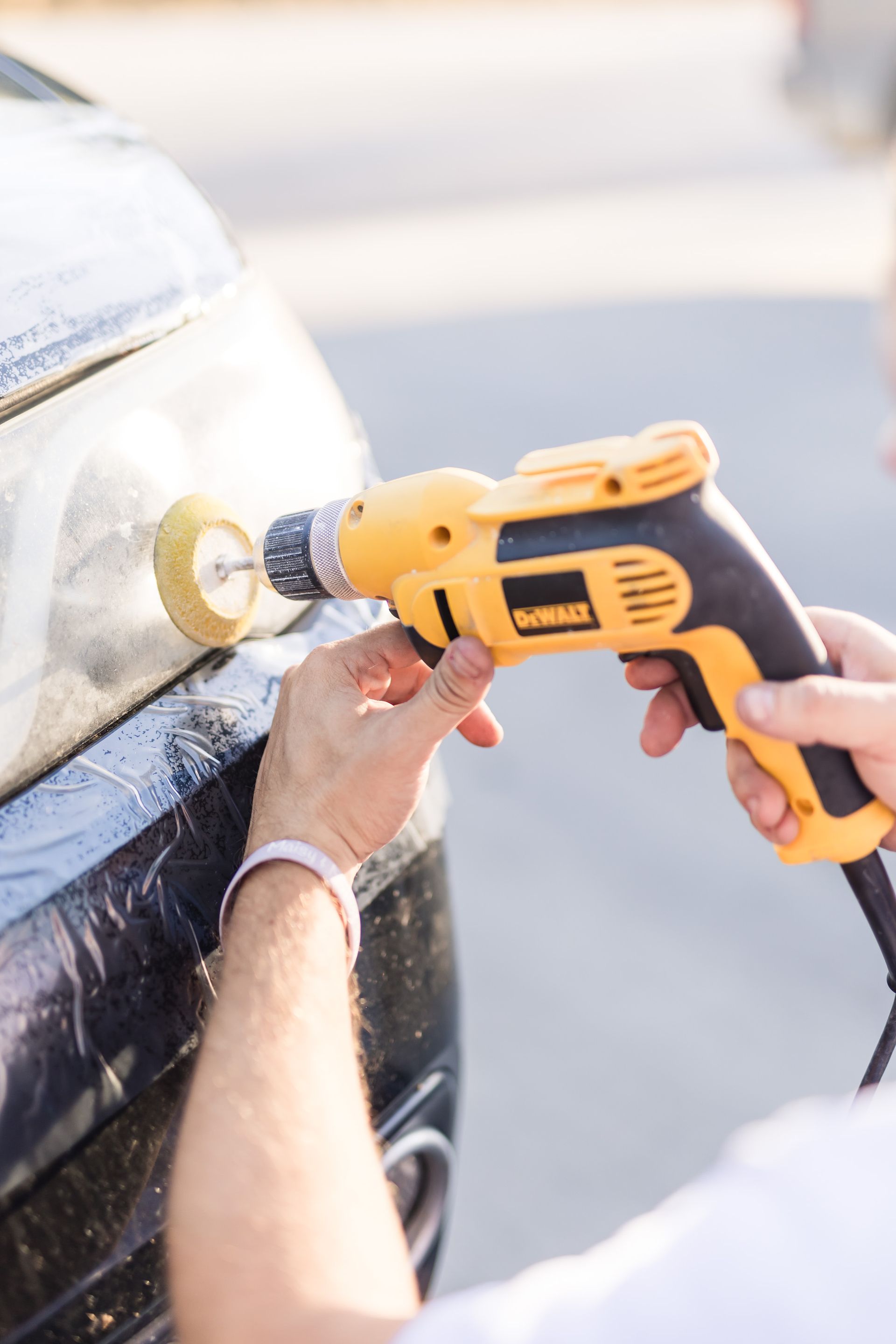 A person polishes a car headlight with a yellow power drill. The car is black and the headlight is cloudy.