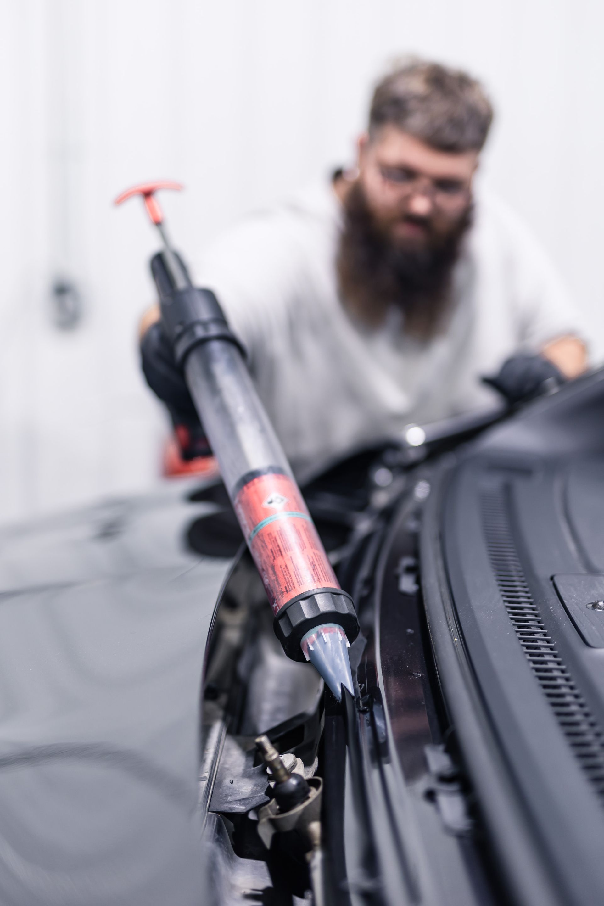 A person, blurred, applying sealant to a black car's trim with a caulking gun. The setting appears to be a workshop.