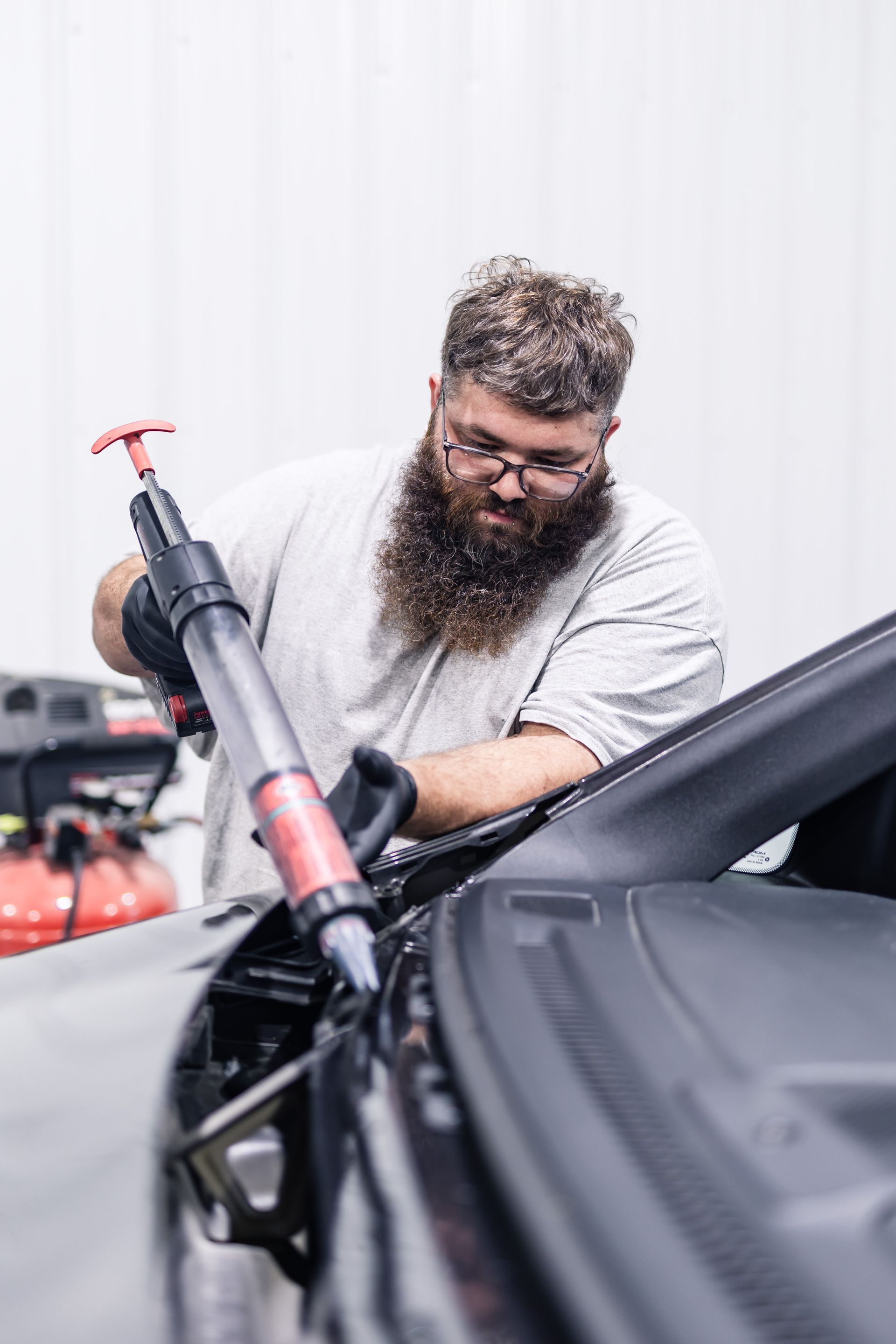A bearded man in gloves uses a fluid extractor on a car in a garage. He is focused, with a neutral expression.