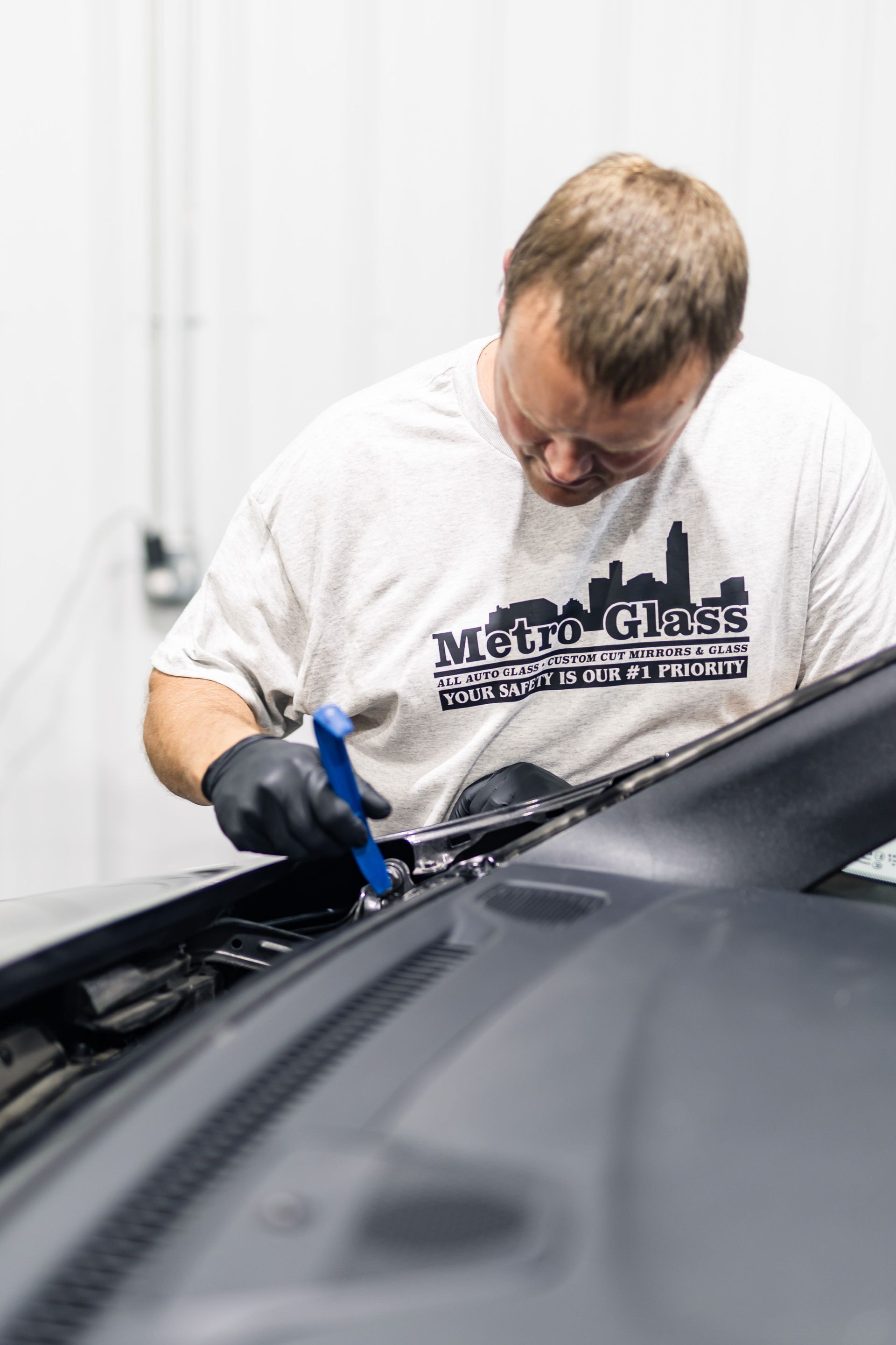 A man in a Metro Glass t-shirt works on a car, using a tool. He wears gloves in a white-walled shop.