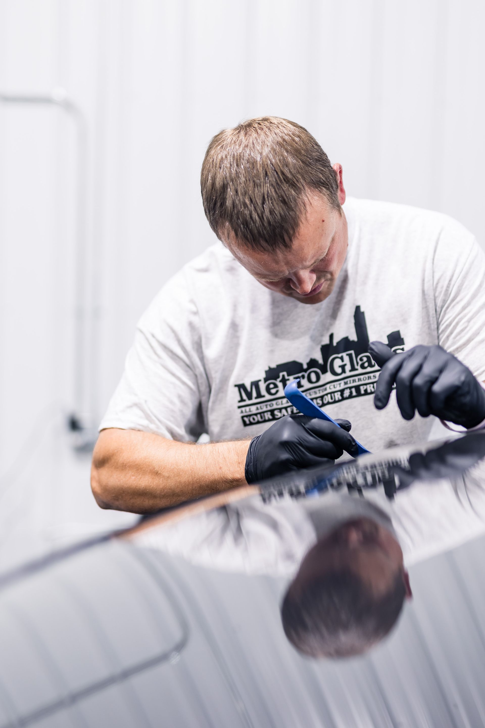 A man wearing a Metro Glass t-shirt and black gloves works on a car's hood, carefully using a tool.