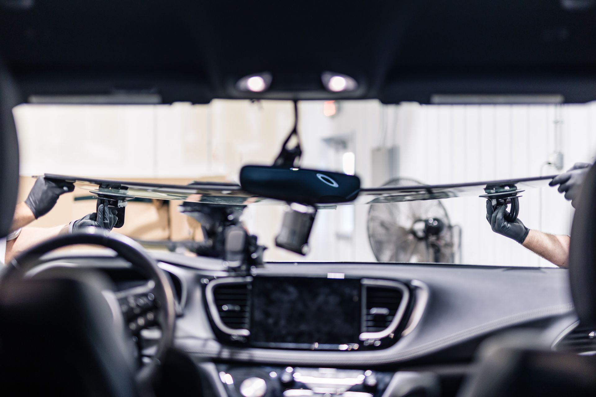 Two technicians install a windshield inside a car, with a dashboard and steering wheel visible.