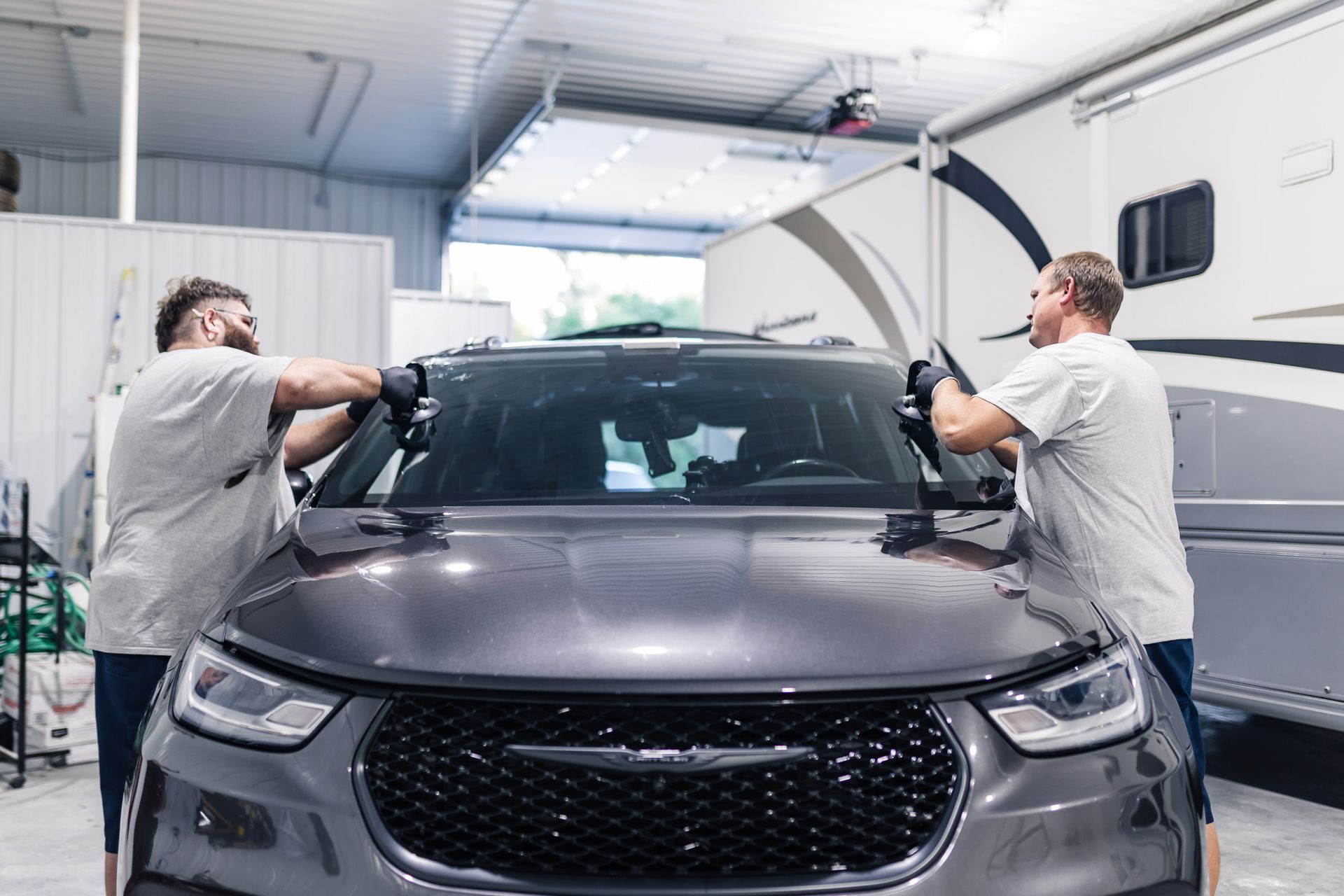 Two men working on the windshield of a dark gray Chrysler Pacifica in a garage.
