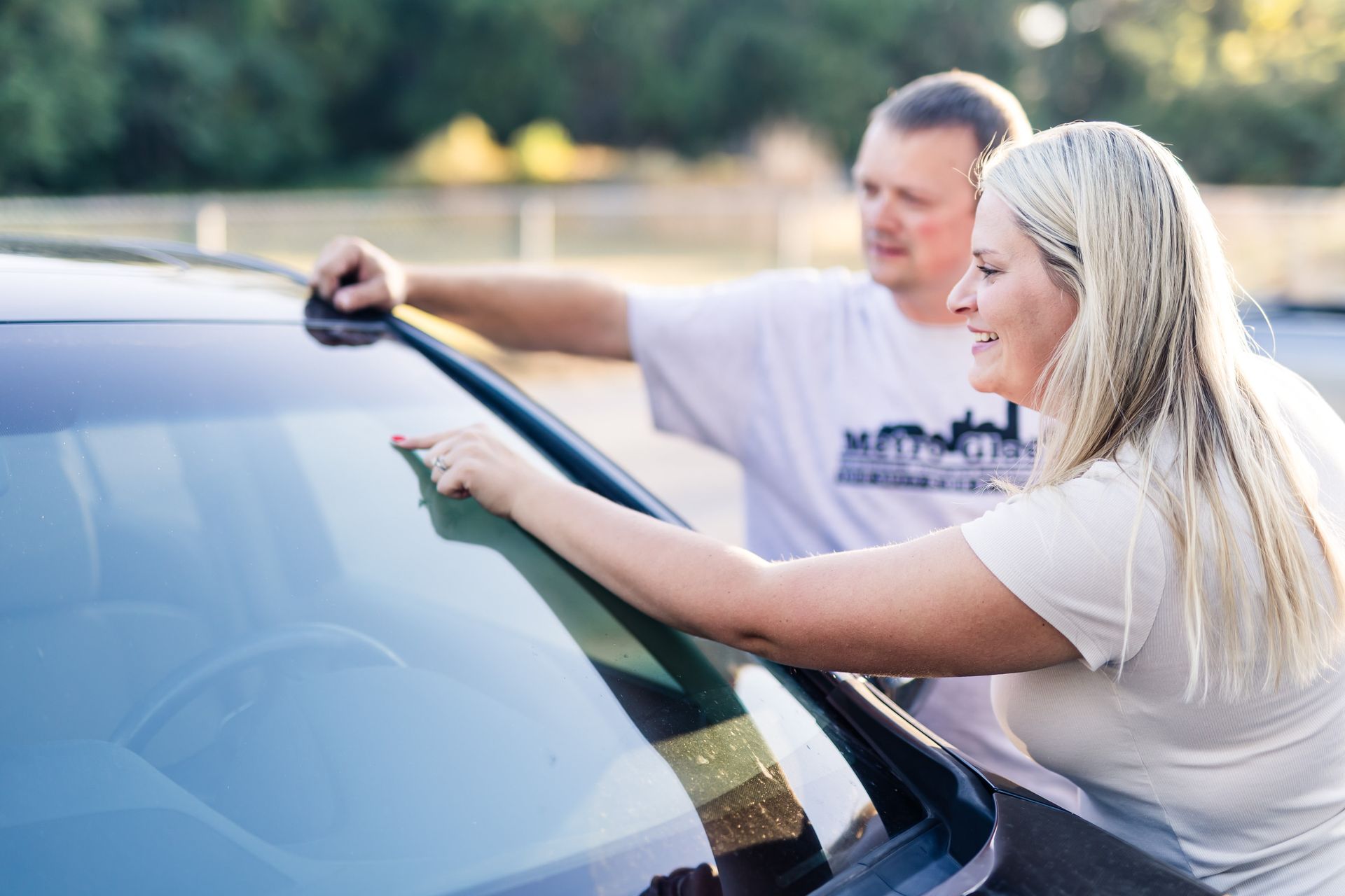 Man and woman examining a car windshield outdoors; woman points, both smile.