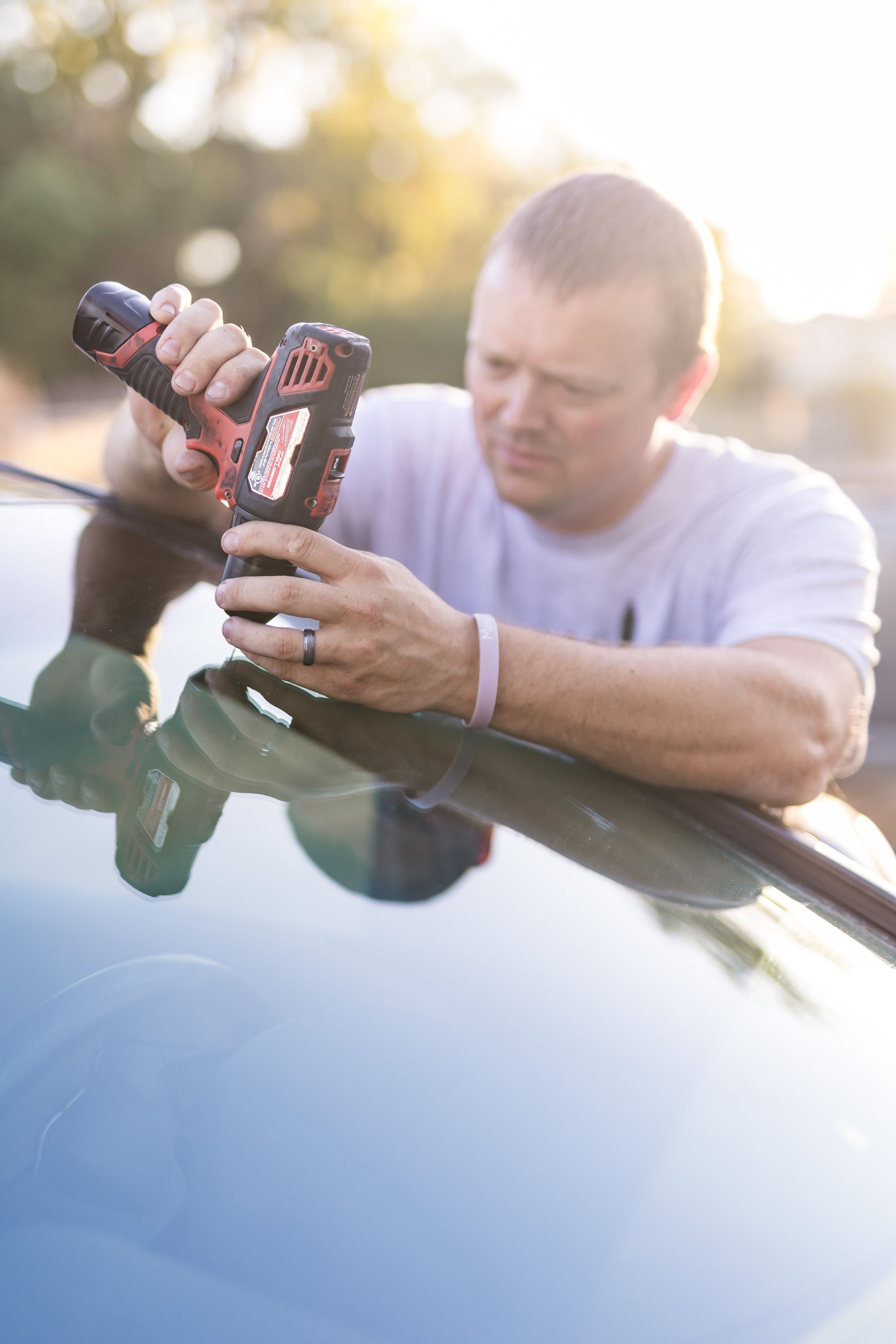 Man using a power drill on a car's windshield, outdoors. He's focused, wearing a white t-shirt. Sunlight is behind him.
