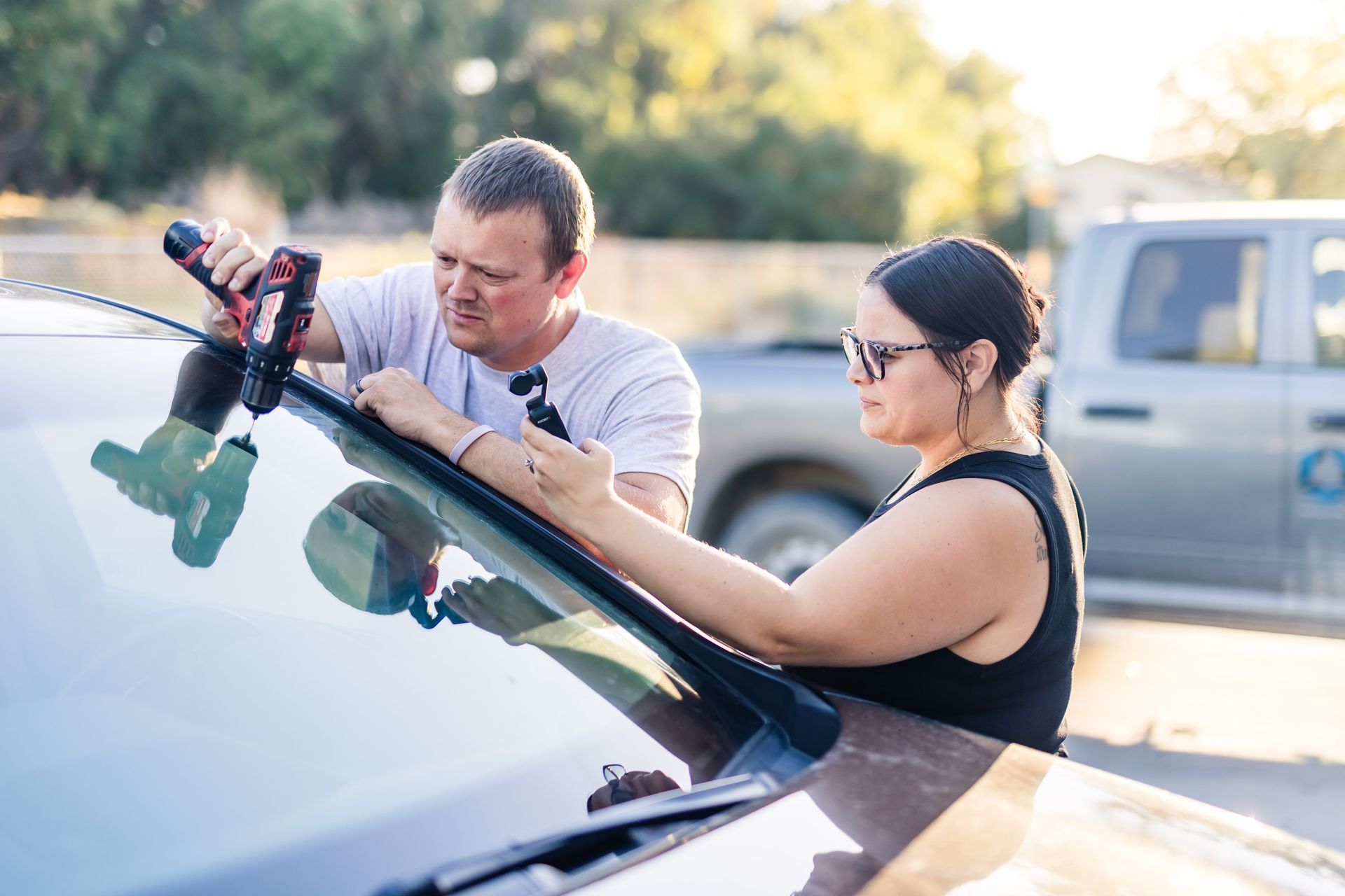 Two people replacing a windshield on a car outdoors. One uses a drill; the other holds the glass.