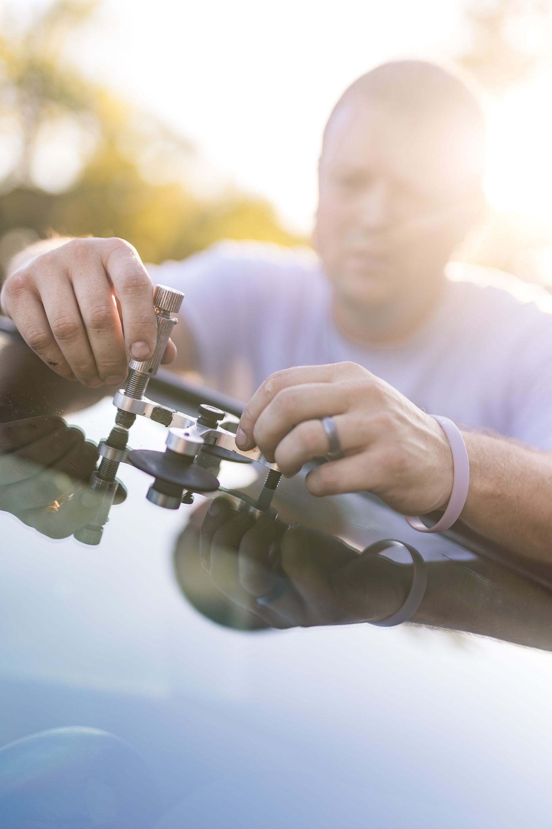 A man repairs a windshield, using tools to fix a crack. He is outdoors, and the sun shines brightly behind him.