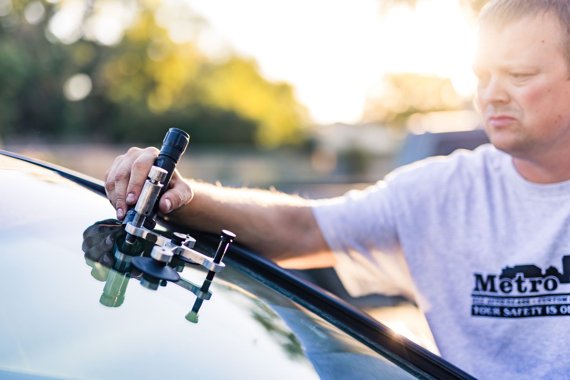 A man in a white shirt repairs a windshield crack with a specialized tool outdoors on a sunny day.