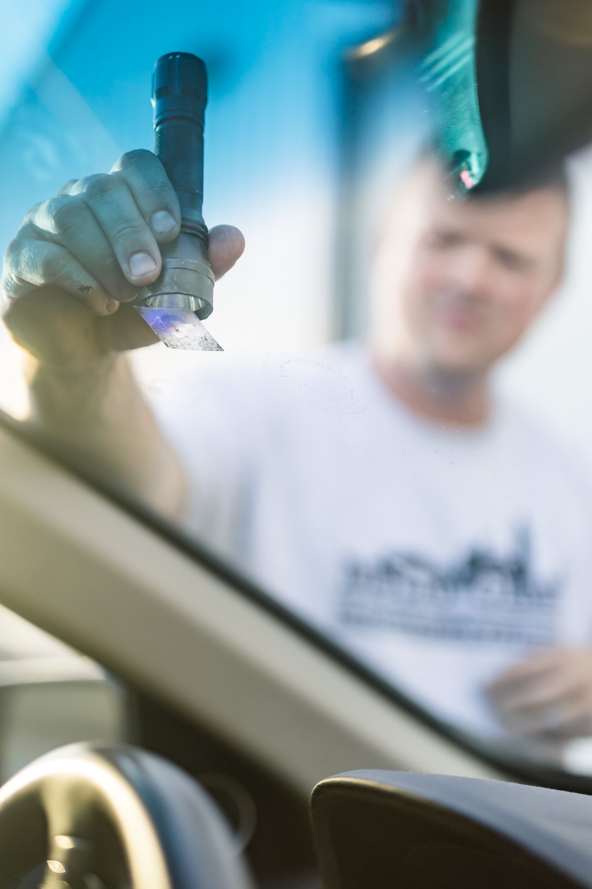 Person shining a flashlight on a car windshield, likely inspecting it. Focus on the flashlight and hand; man is blurry in the background.
