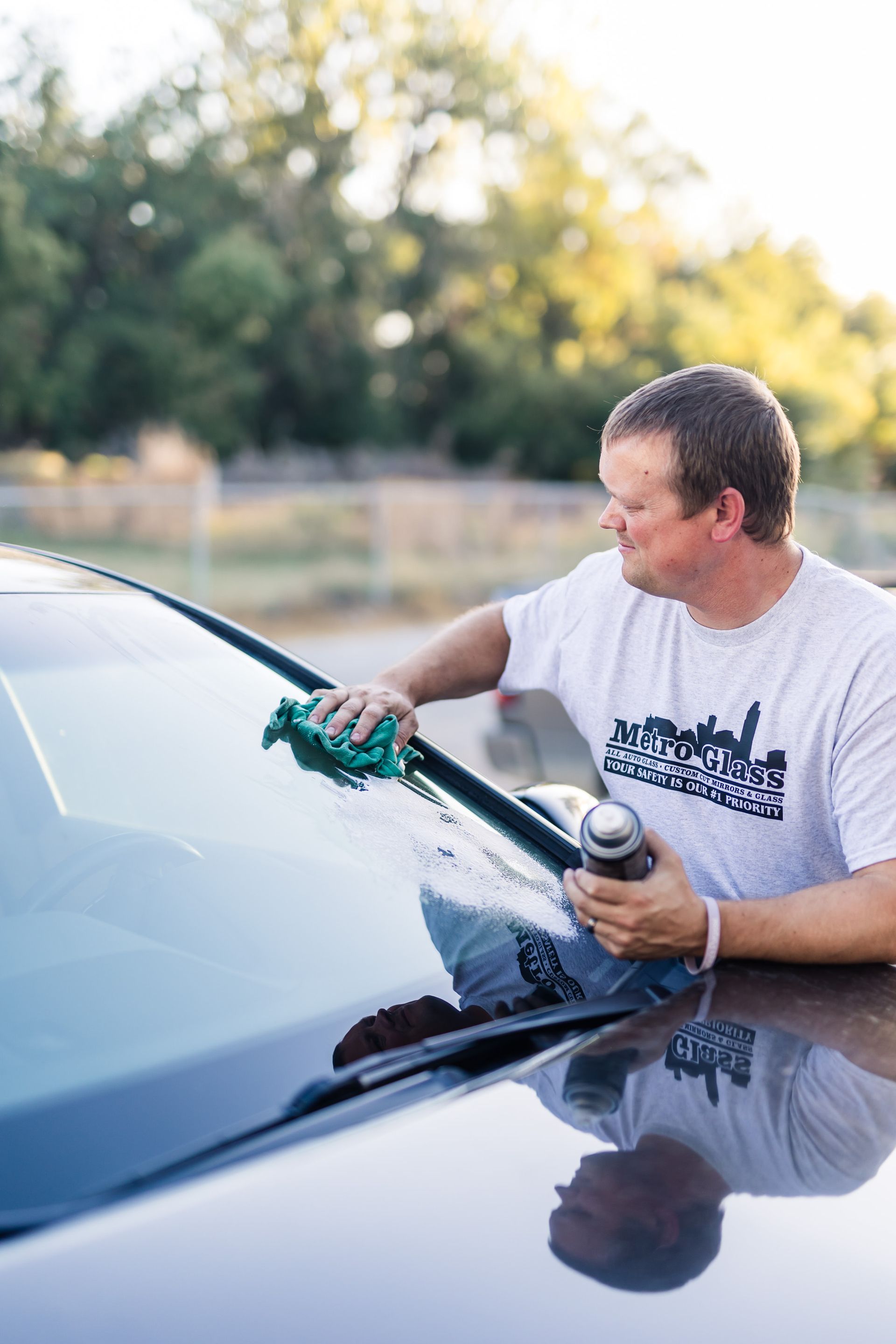 Man cleaning a car windshield outdoors with a spray bottle and a green cloth. He is wearing a white shirt and appears focused.
