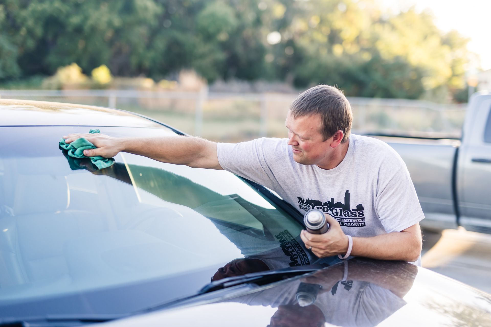 Man cleaning a car windshield with a cloth and spray bottle outdoors. He is wearing a white t-shirt, and the setting is a parking lot.