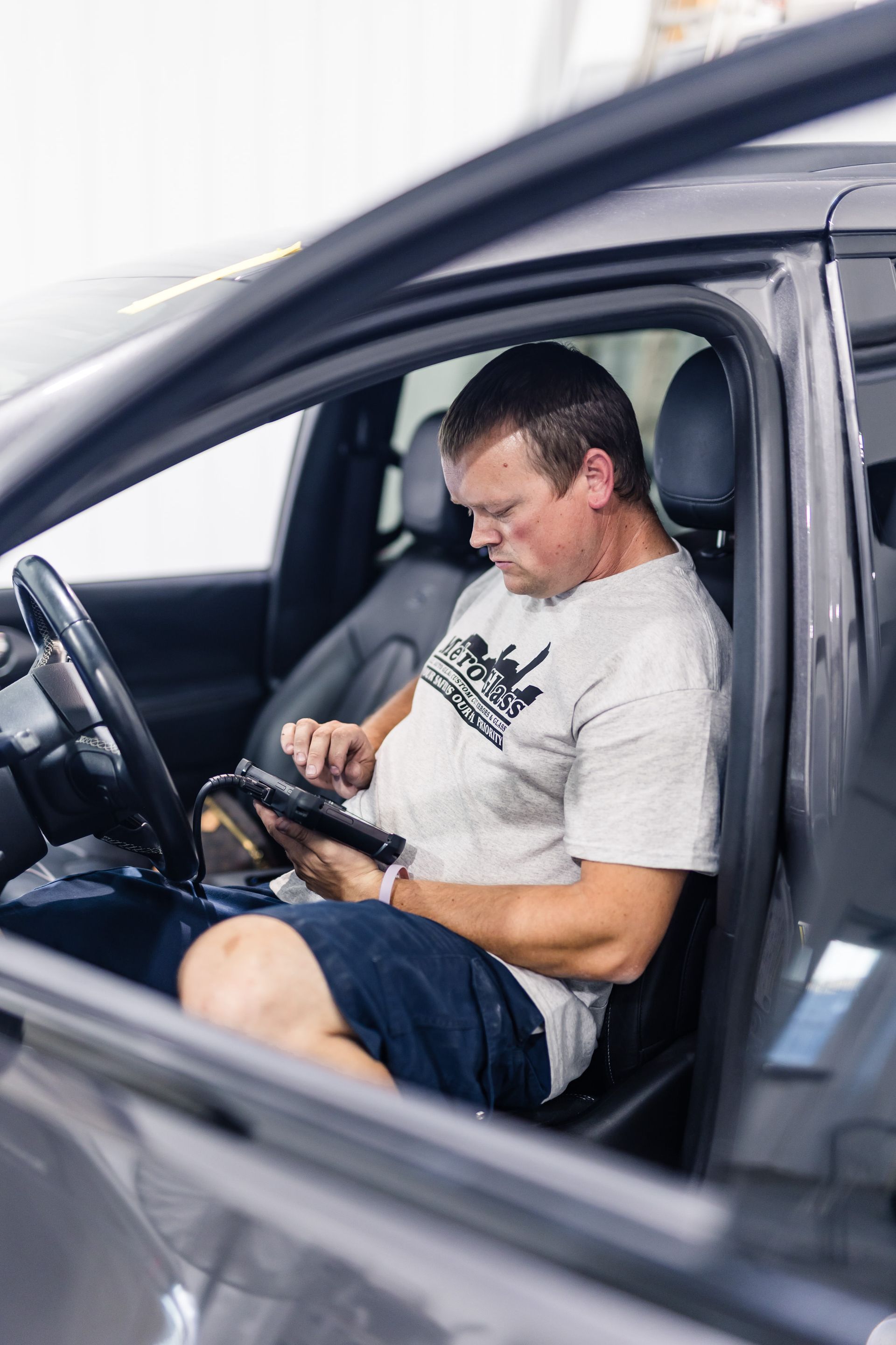 A man in a car uses a tablet. He wears a gray shirt and blue shorts, seated in a vehicle with the door open, likely for diagnostics.