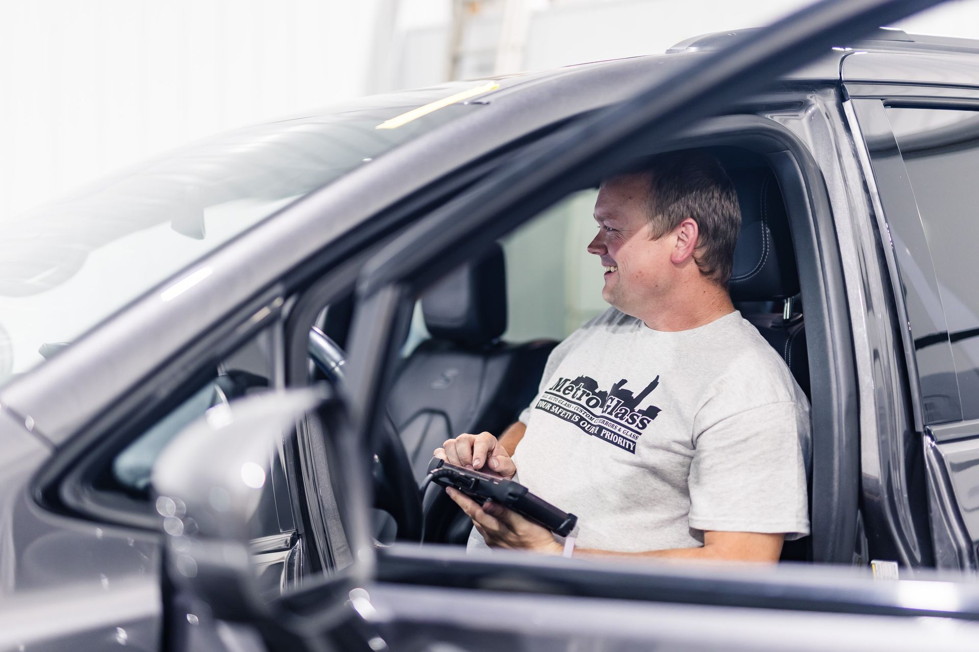 Man in a car, smiling and holding a device. He is wearing a light t-shirt and sitting in a black interior.