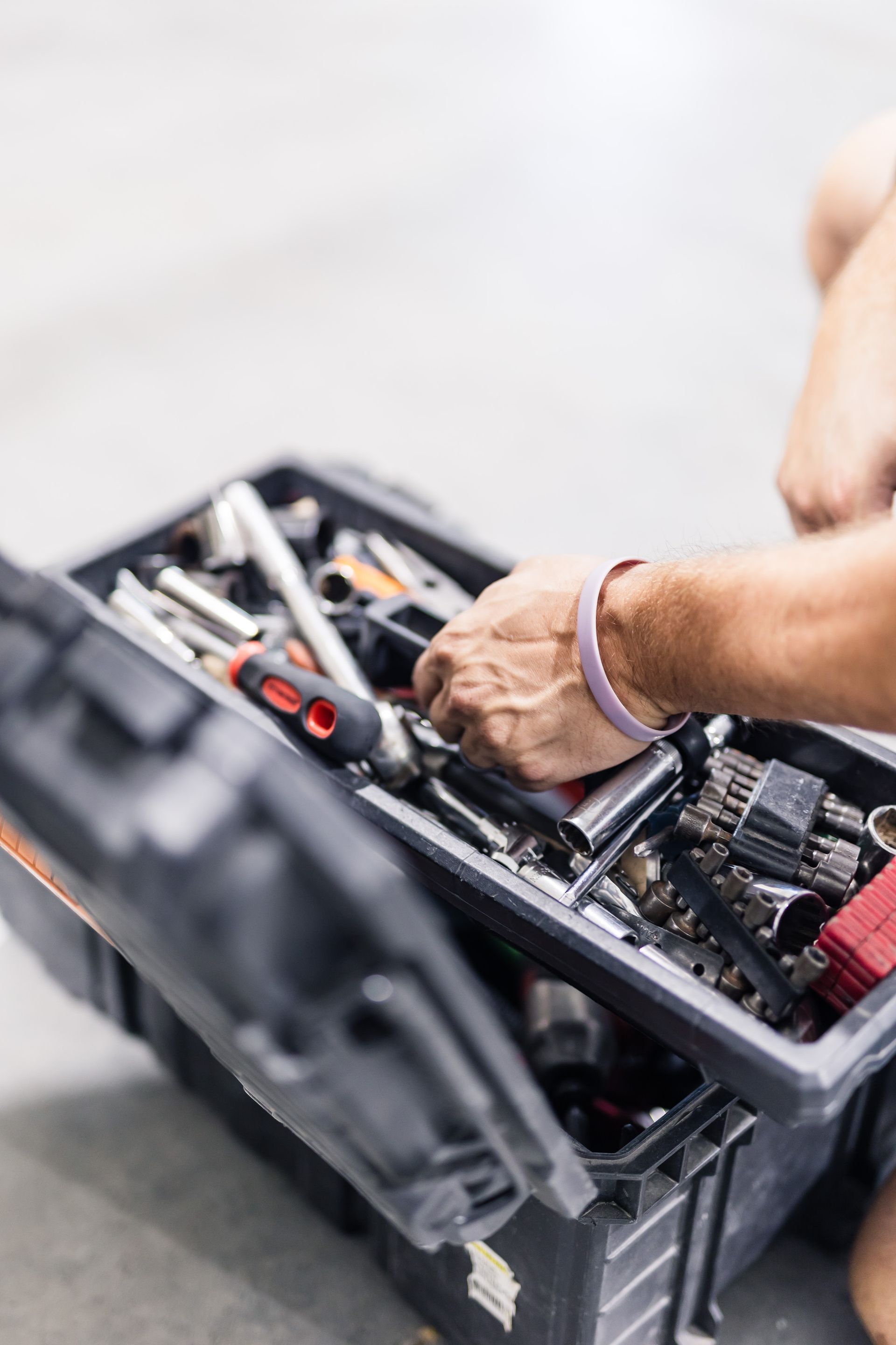 A person's hand reaching into an open black toolbox filled with various tools, in a bright, concrete setting.
