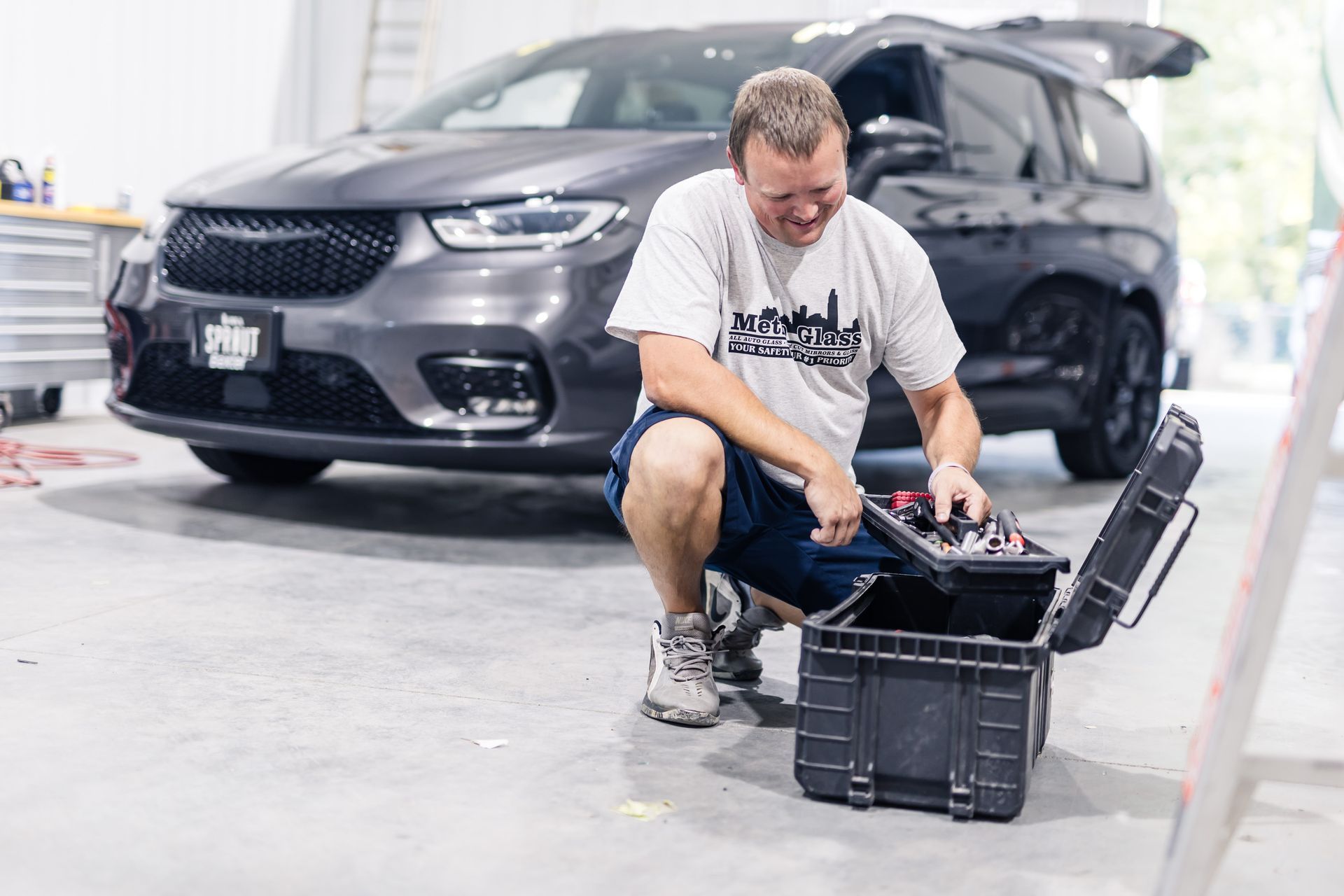 A man kneels, opening a toolbox in front of a gray minivan inside a garage. He smiles, wearing a t-shirt, shorts, and sneakers.