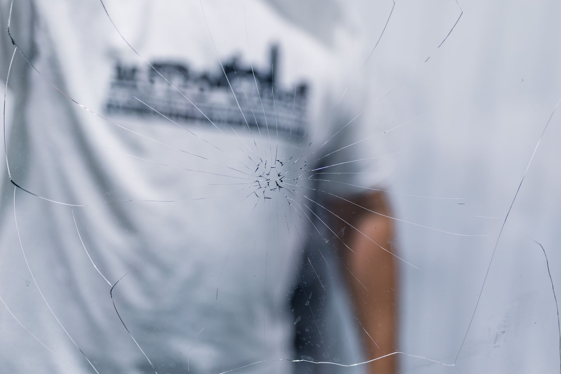 Spiderweb in focus, partially obscuring a person wearing a light-colored shirt with a graphic on the back, against a blurred white background.