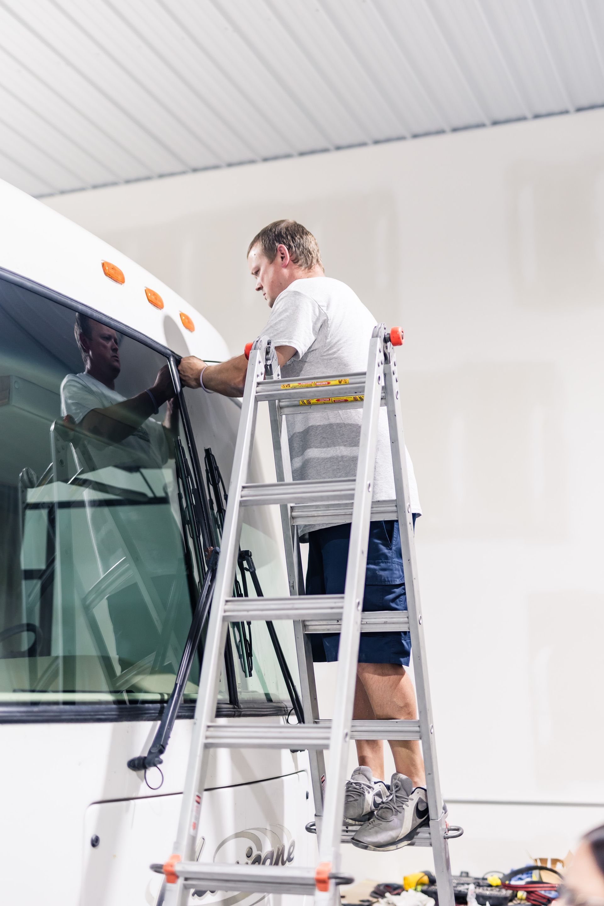 A man on a ladder is working on the windshield of a white RV in a well-lit garage.