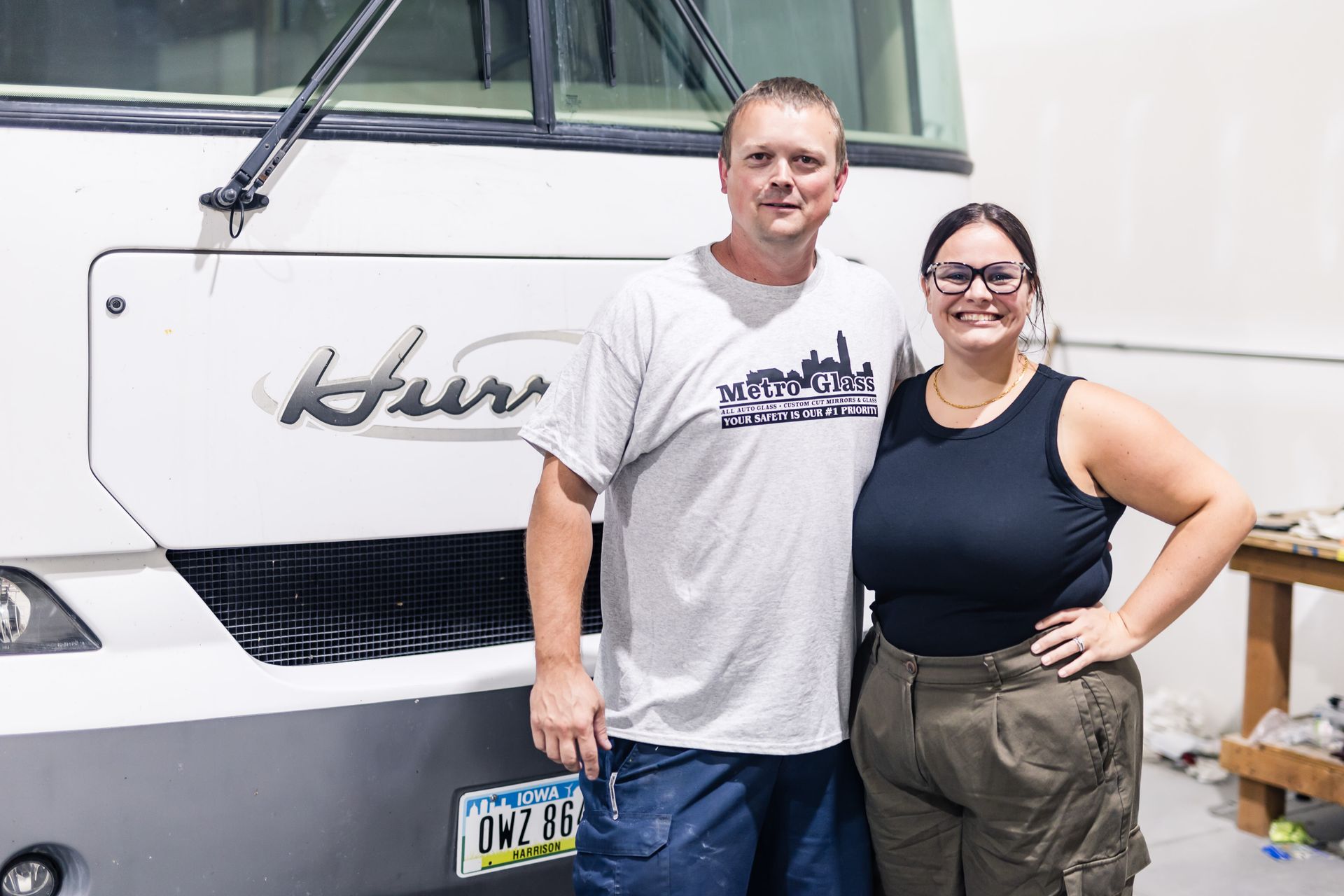 A man and woman pose in front of a white RV. The man wears a gray t-shirt, and the woman wears a black tank top. They smile.