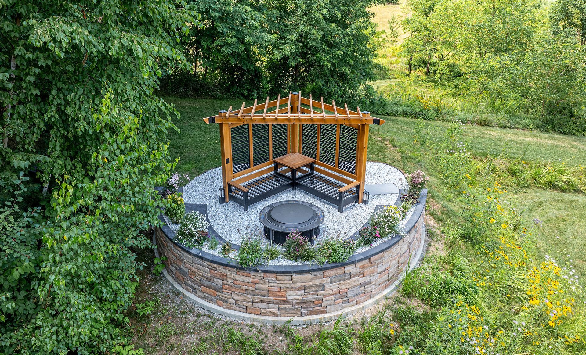 Gazebo with seating around a fire pit on a circular stone patio, surrounded by landscaping and trees.