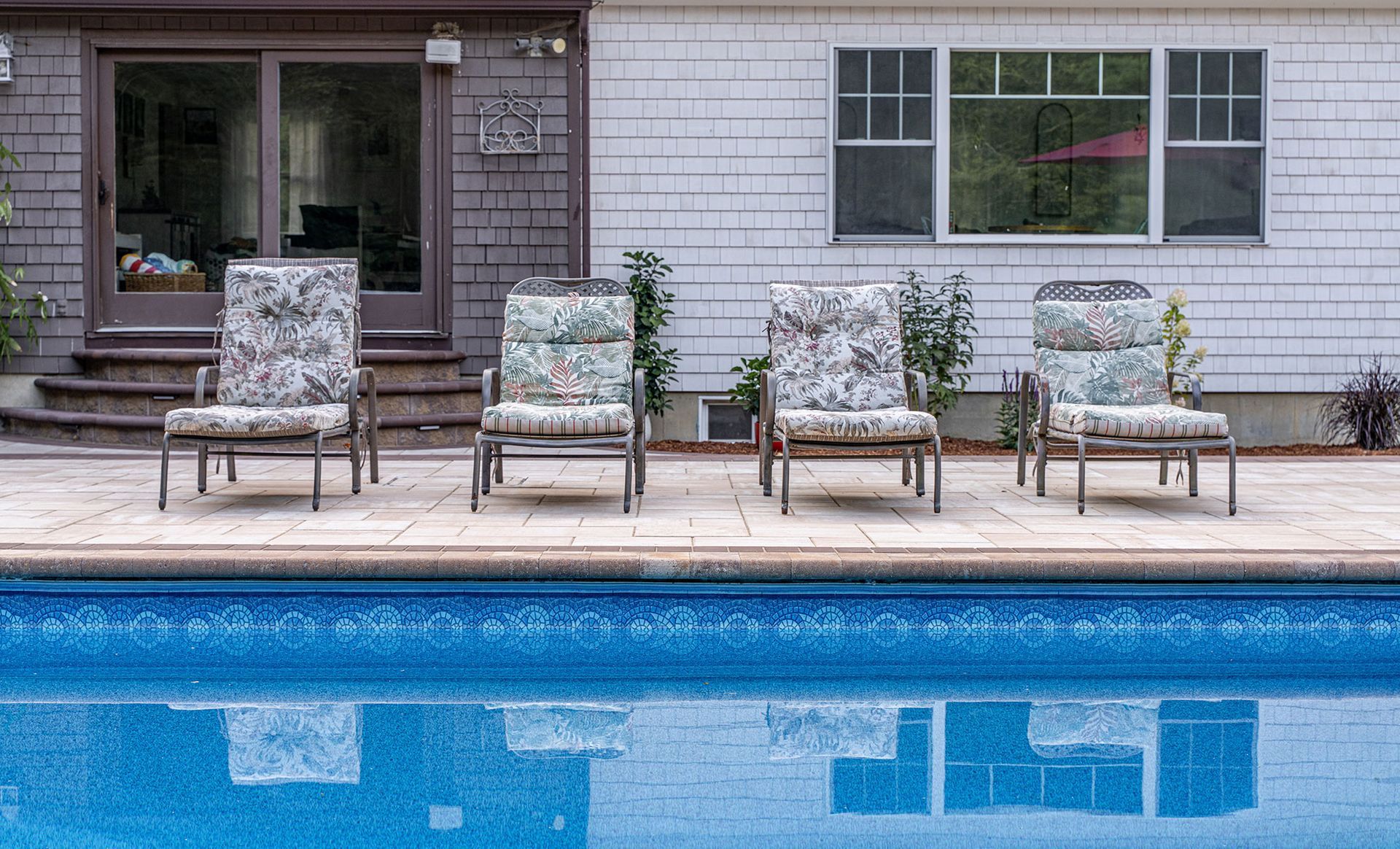 Poolside seating: four patterned chairs line a pool's edge, reflecting in the blue water, with a house in the background.