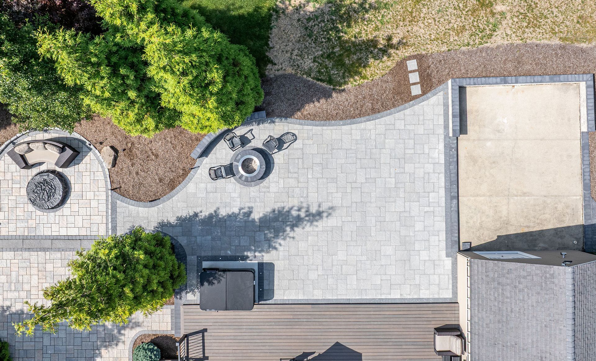 Overhead view of a backyard patio with gray pavers, fire pits, wood deck, and landscaping.