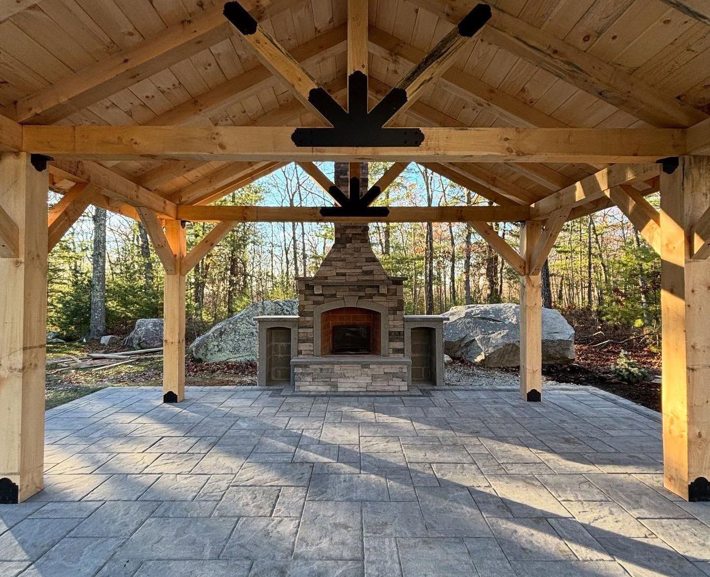 Wooden pavilion with a stone fireplace, on a gray stone patio. Forest in the background.